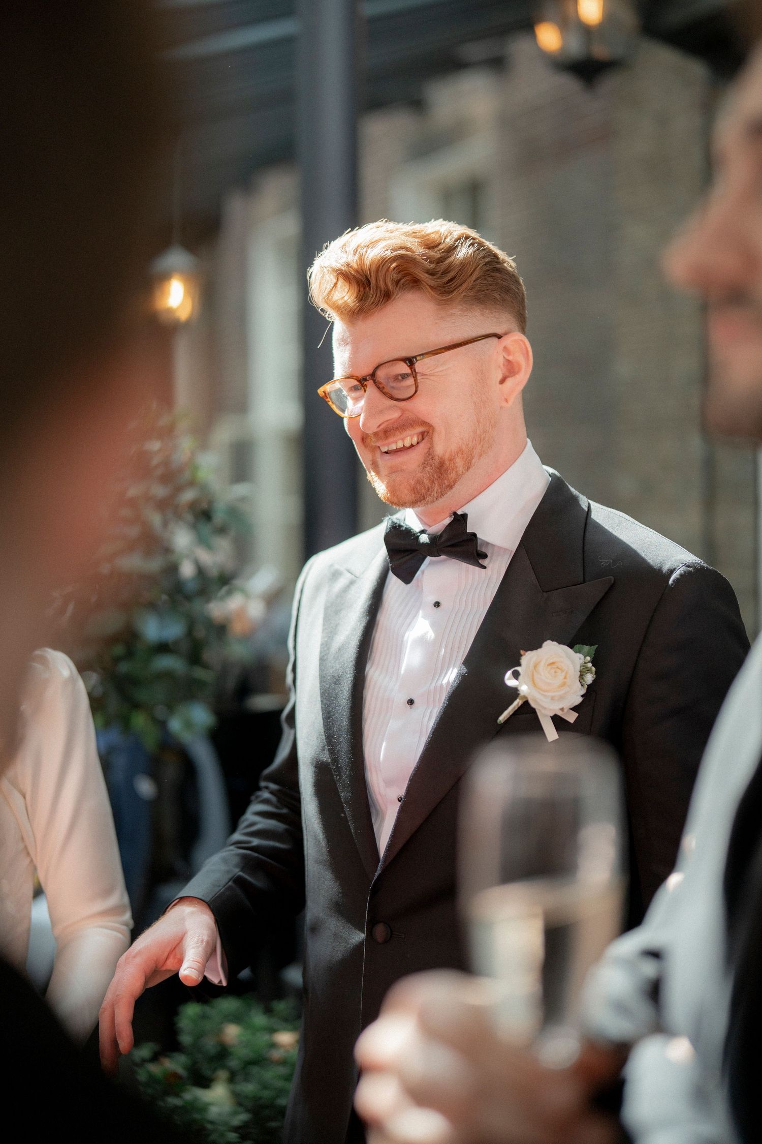 Smiling red-haired groom in tuxedo with boutonniere enjoying outdoor wedding reception with champagne guests.