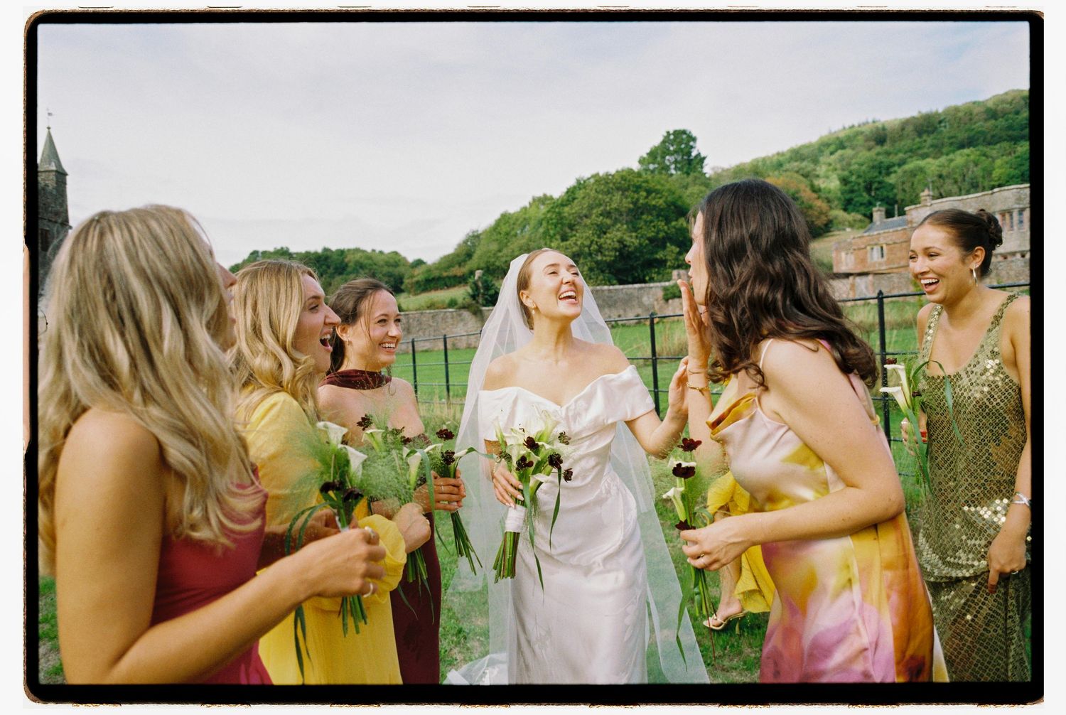 A joyful bride in a white gown laughs with her bridesmaids in colorful mismatched dresses outdoors.