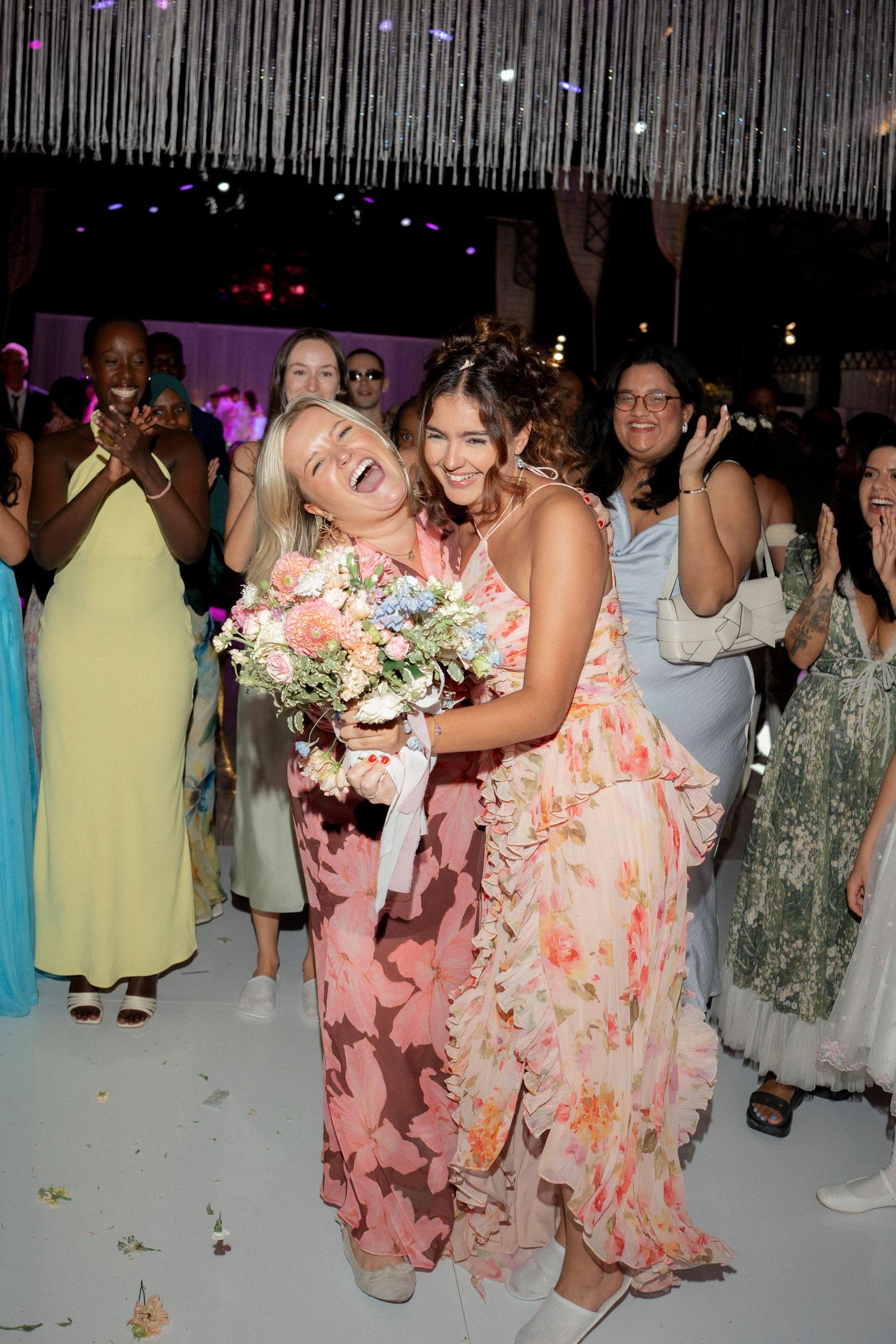 Two women in floral dresses laugh joyfully after catching the bouquet at a wedding.