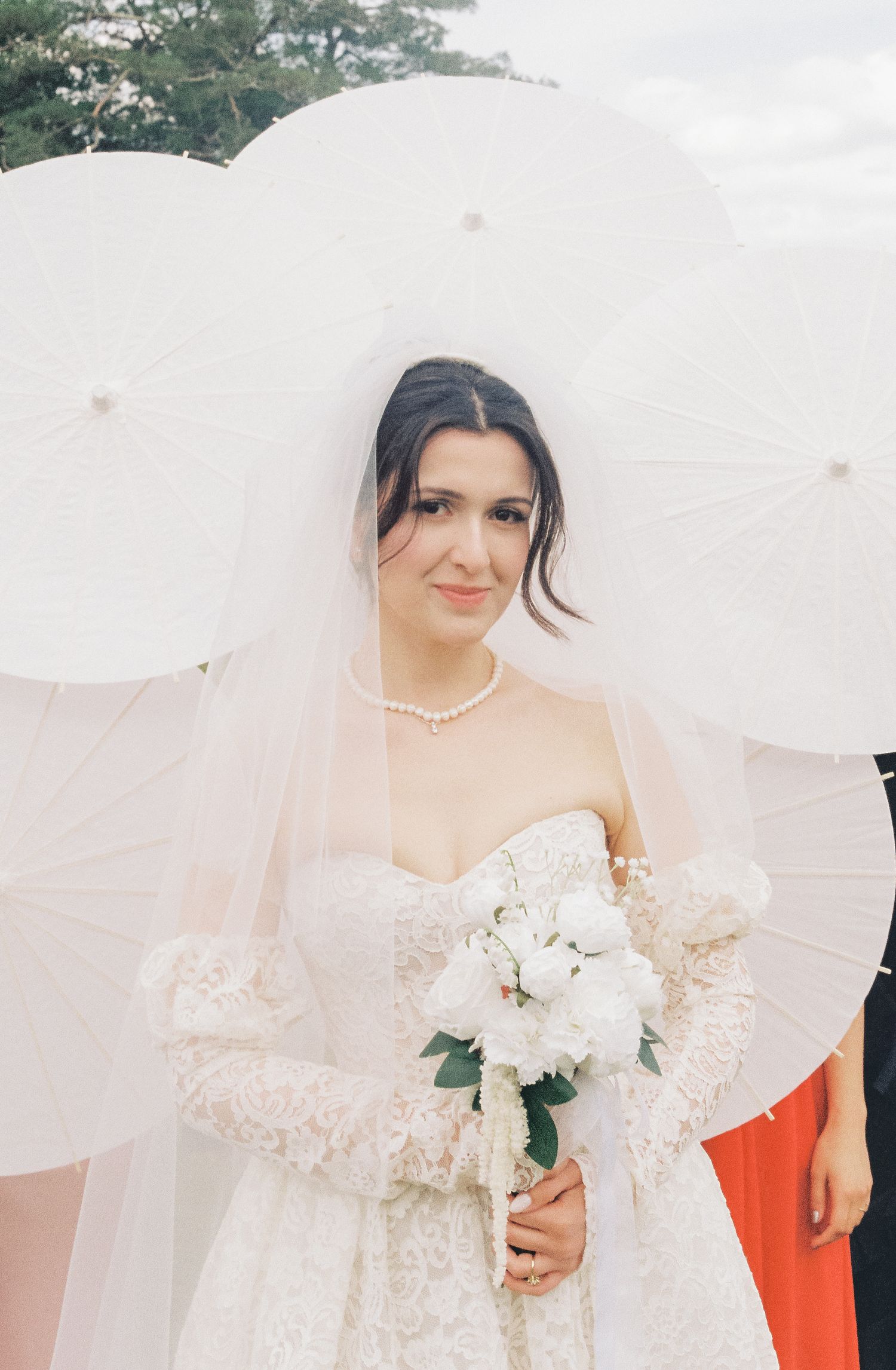 Bride in a flowing white lace dress and veil holds a bouquet of white flowers outdoors.