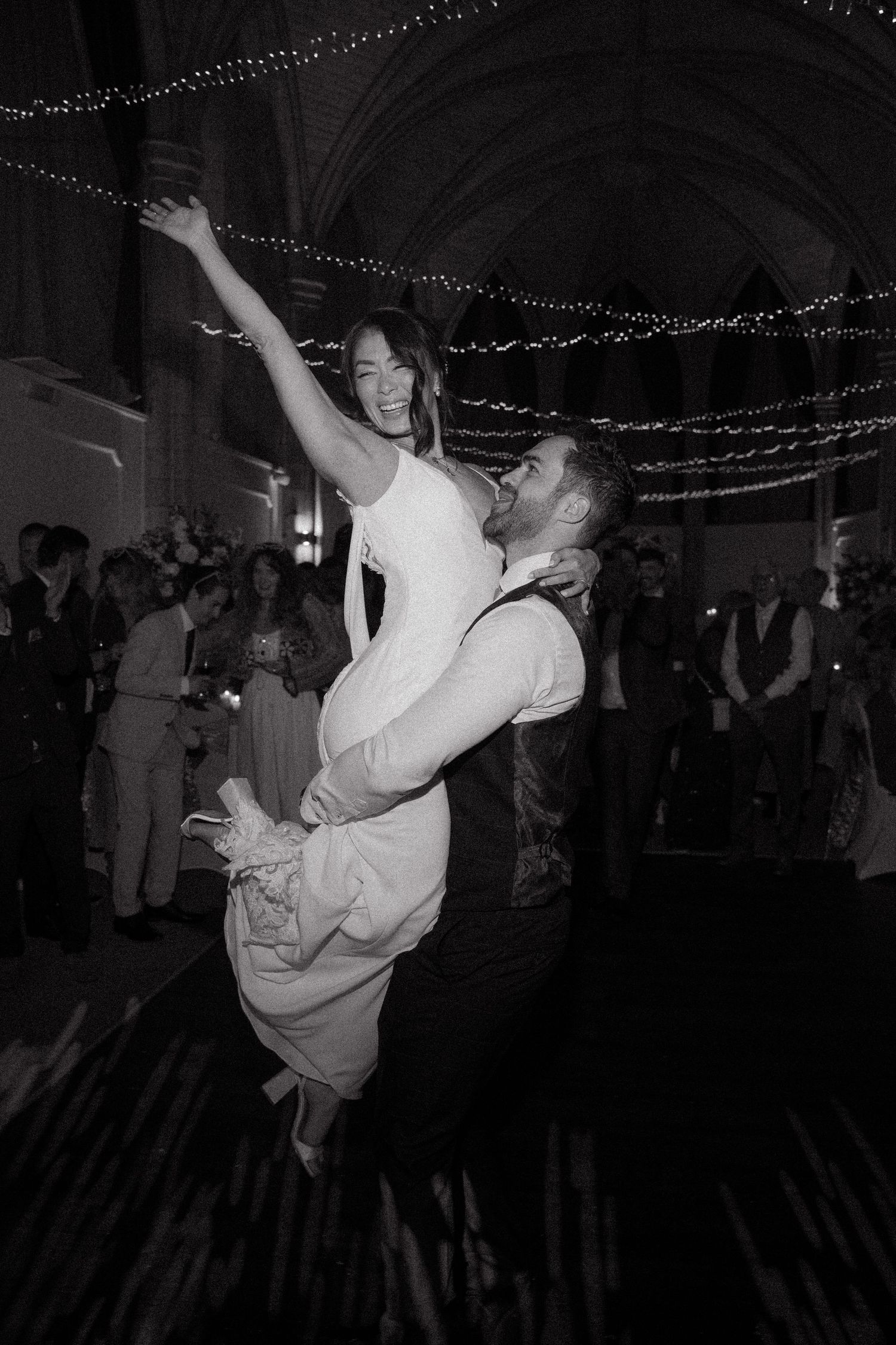 A couple performs an energetic dance lift move at a dimly lit reception venue with string lights overhead.