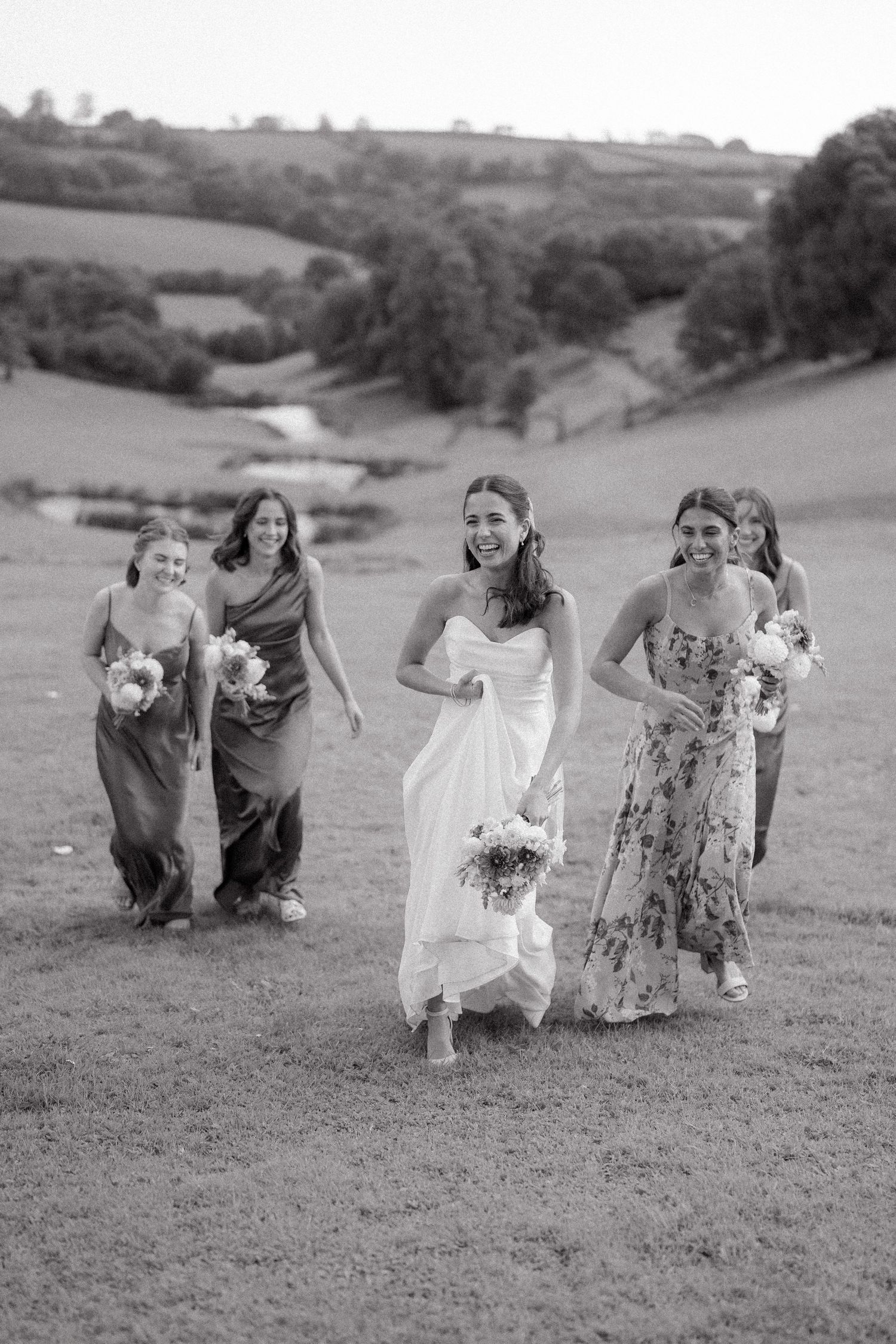 Wedding party members run playfully across a grassy hillside in black and white.