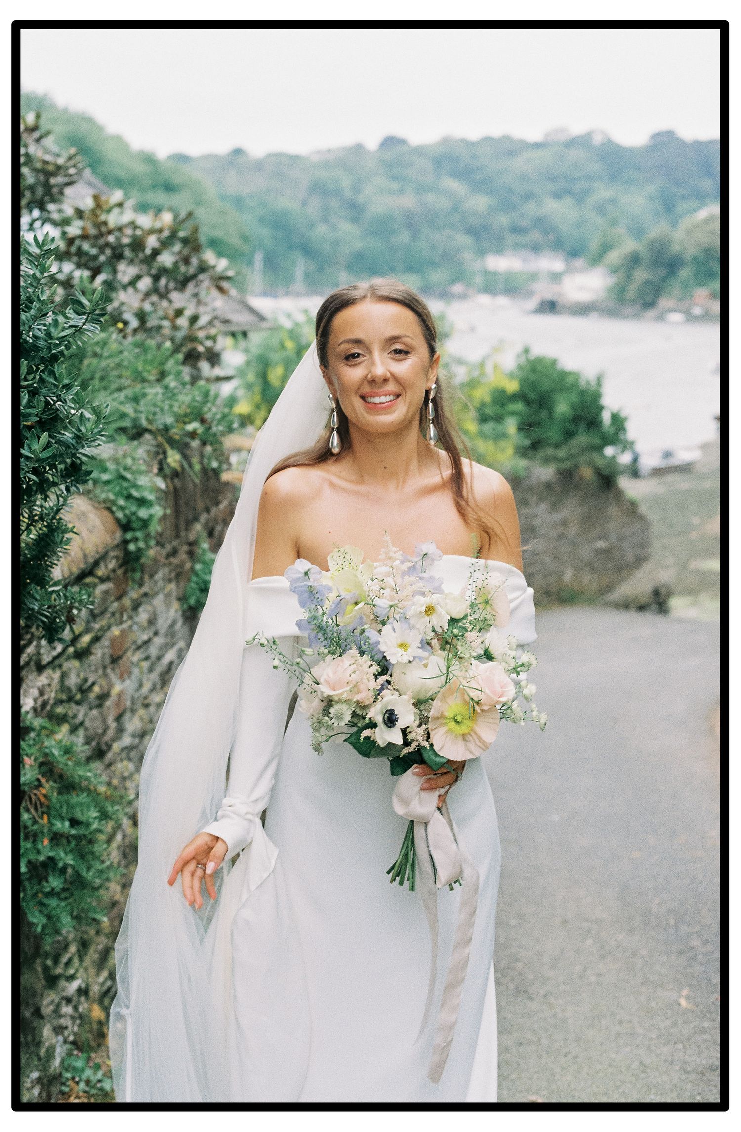 Elegant bride in off-shoulder dress holding a pastel bouquet against water view.