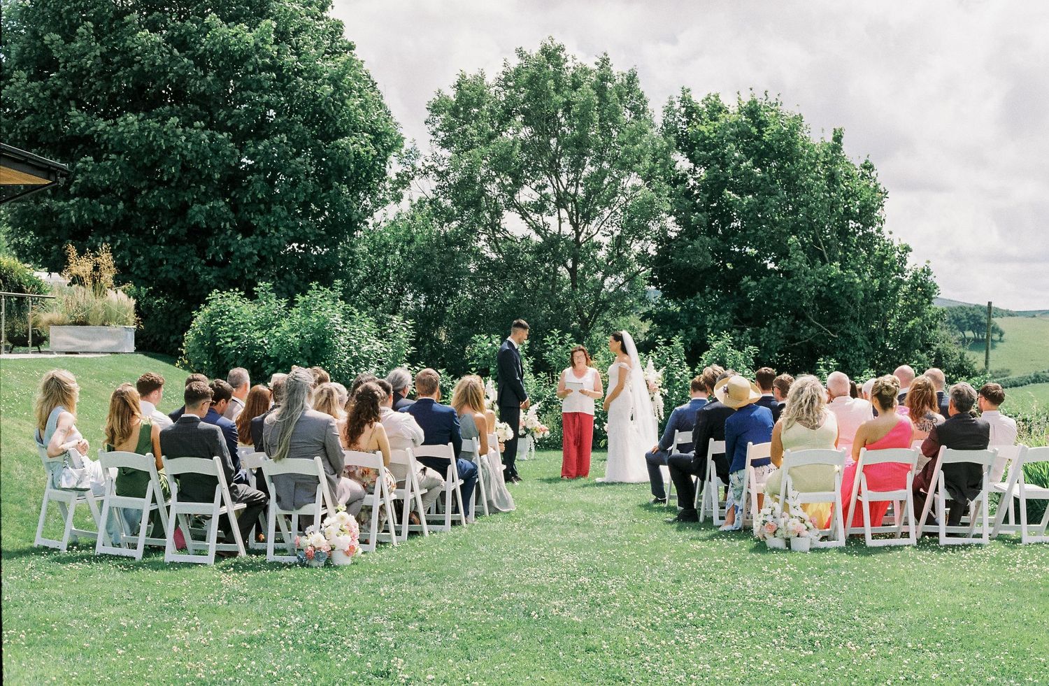 Panoramic view of wedding guests seated in white chairs at an outdoor ceremony venue.