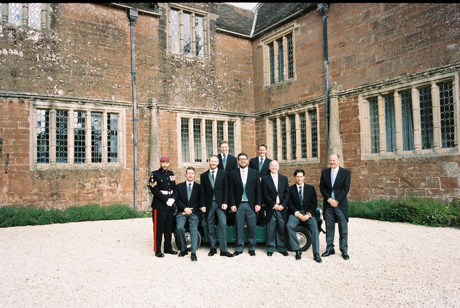 Group of men in dark suits and one man in military dress uniform posing on a gravel courtyard in front of a historic red brick manor house.