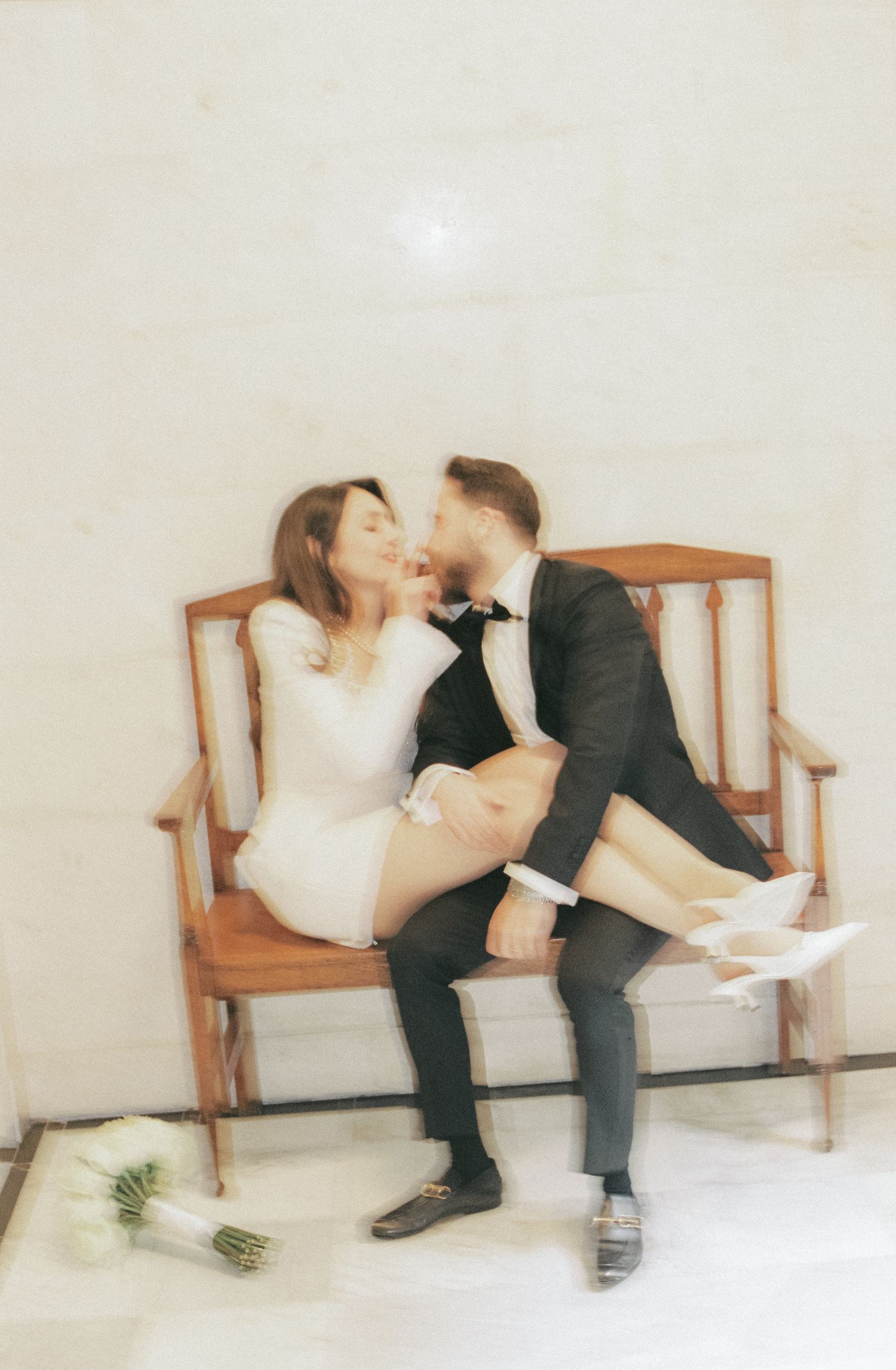 A couple in formal attire shares a romantic moment while sitting together on a wooden bench in a minimalist setting.