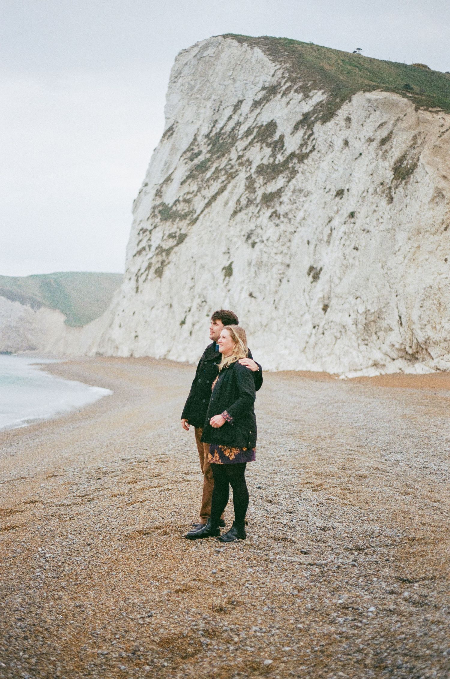 Two people embrace on a pebble beach with dramatic white chalk cliffs rising in the background.