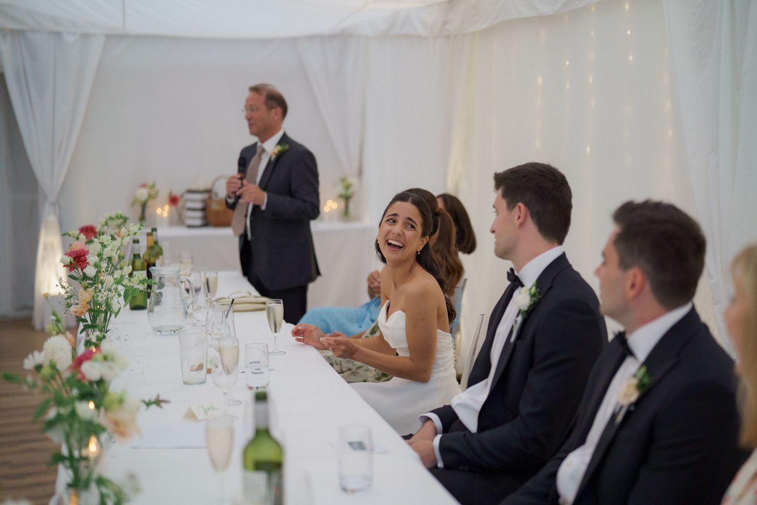 Wedding guests seated at elegant reception tables laugh and smile during dinner celebration.