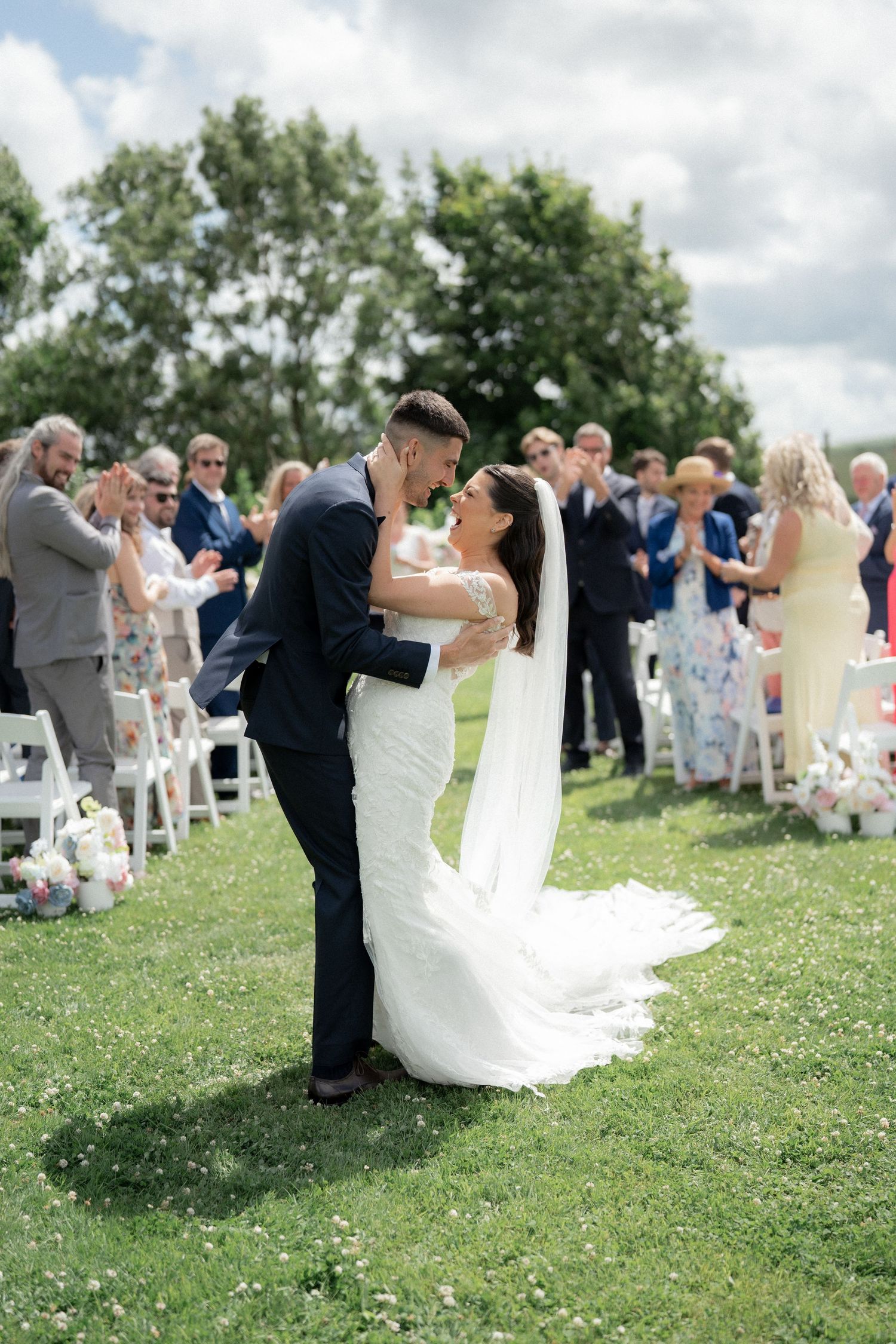 A couple shares a romantic dip kiss on a grassy hillside during their outdoor wedding ceremony while guests look on.