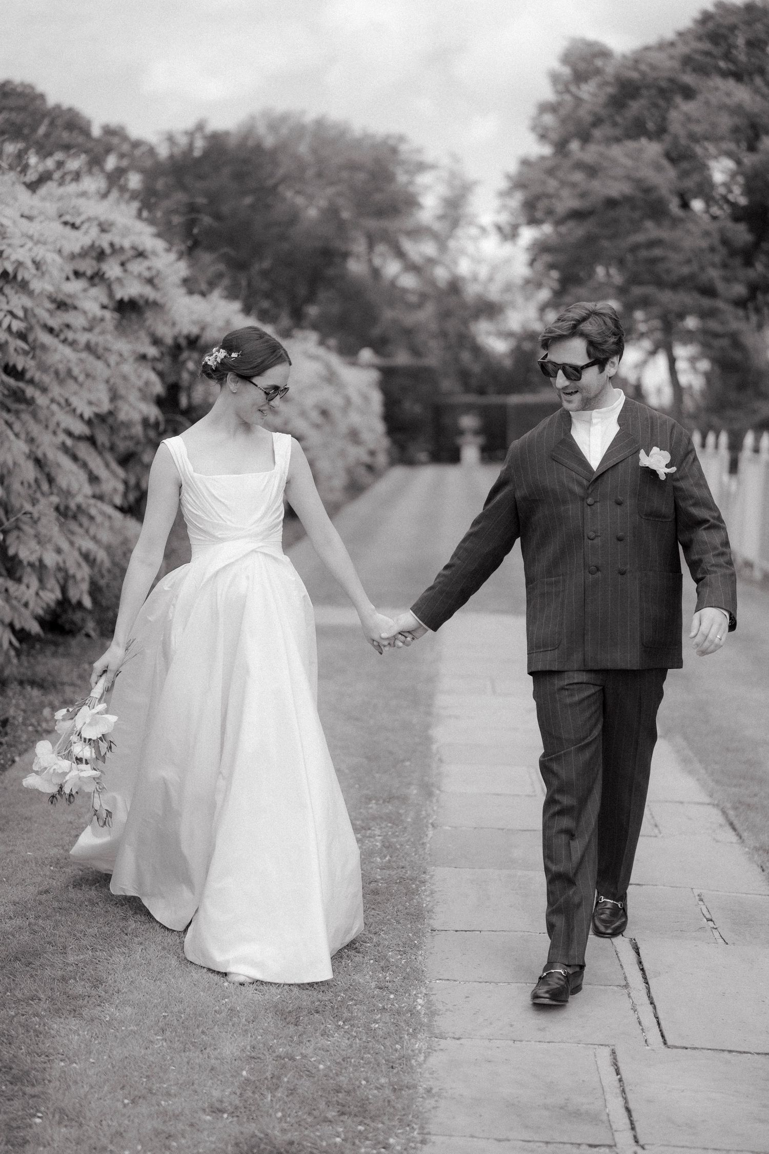 Bride in an elegant white gown and groom in a pinstripe suit and sunglasses holding hands while walking down a garden path.
