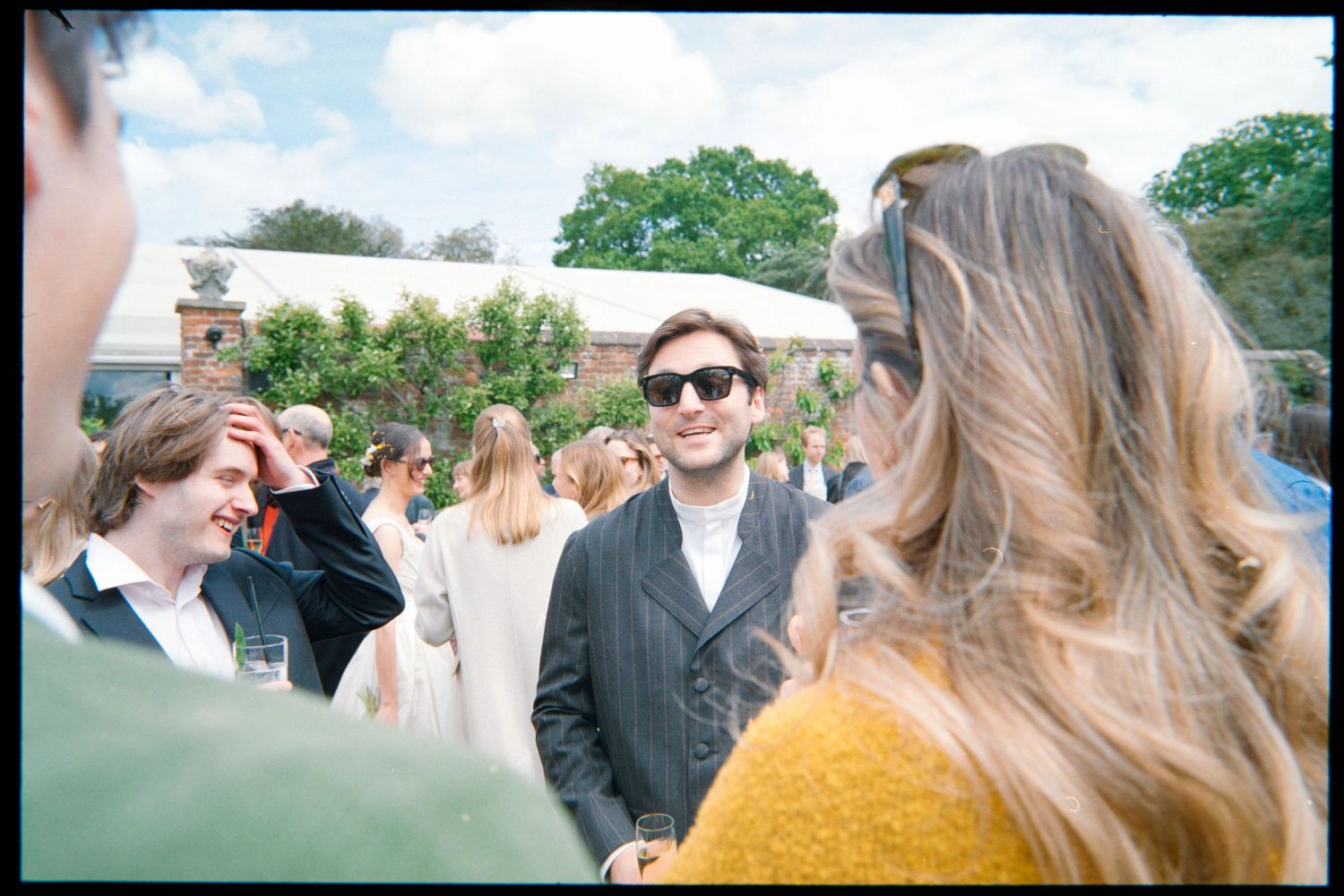 Wedding guests mingle on a sunny lawn, with a smiling man in sunglasses and a tweed jacket facing the camera.