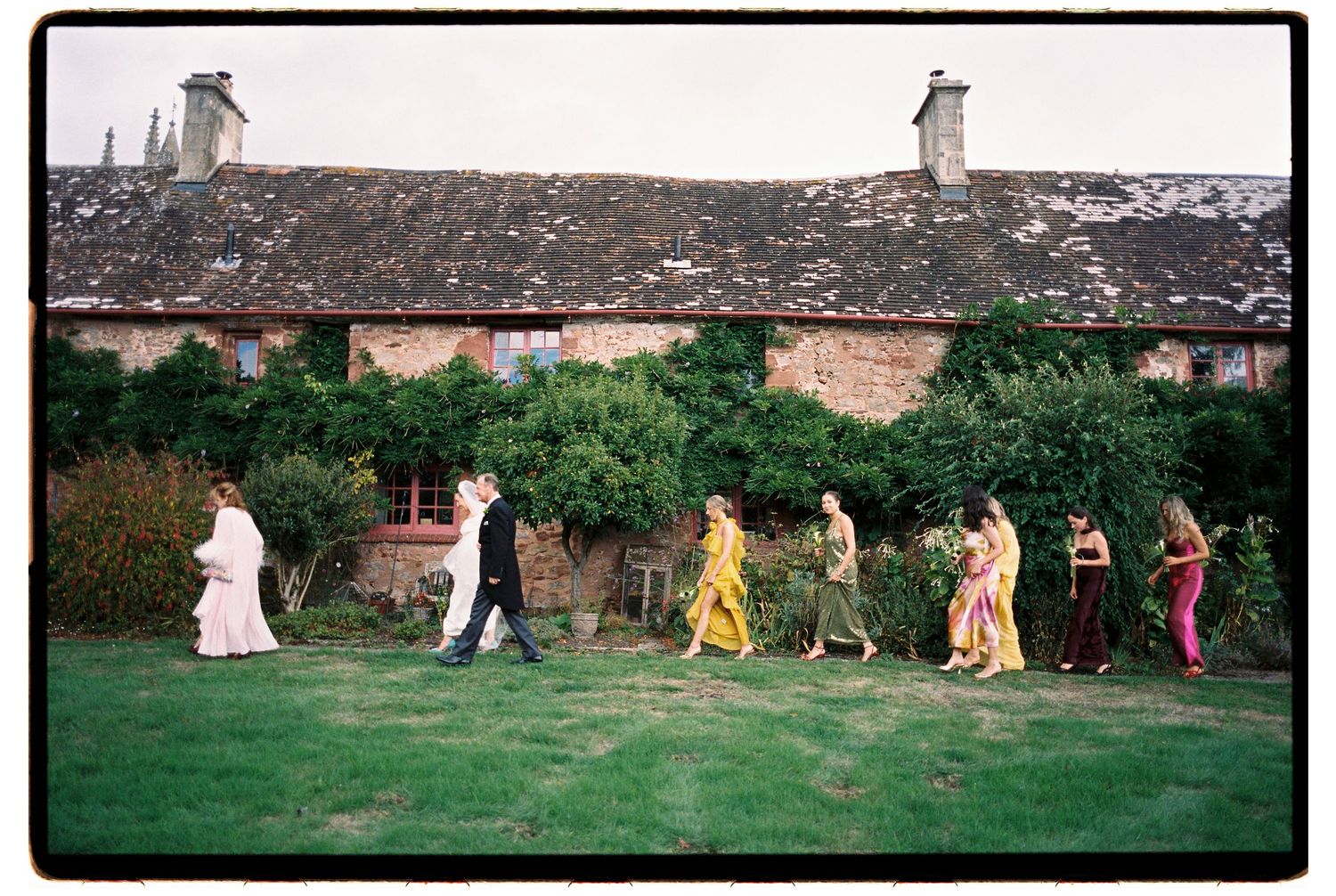 Wedding party walking across a green lawn in front of a rustic stone cottage, bride and groom embracing nearby.