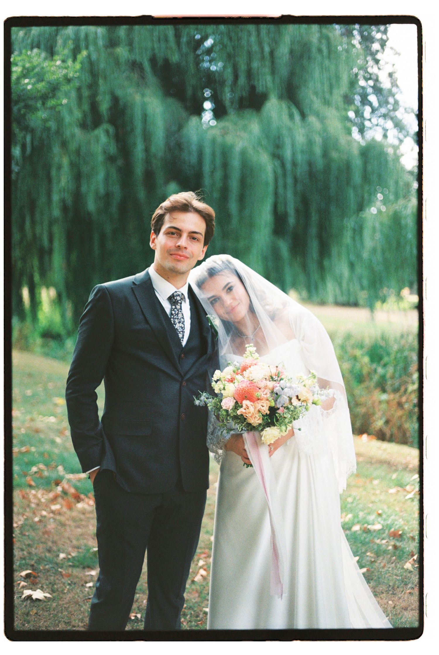 Bride and groom pose together outdoors under a weeping willow tree, bride holding a pastel bouquet.