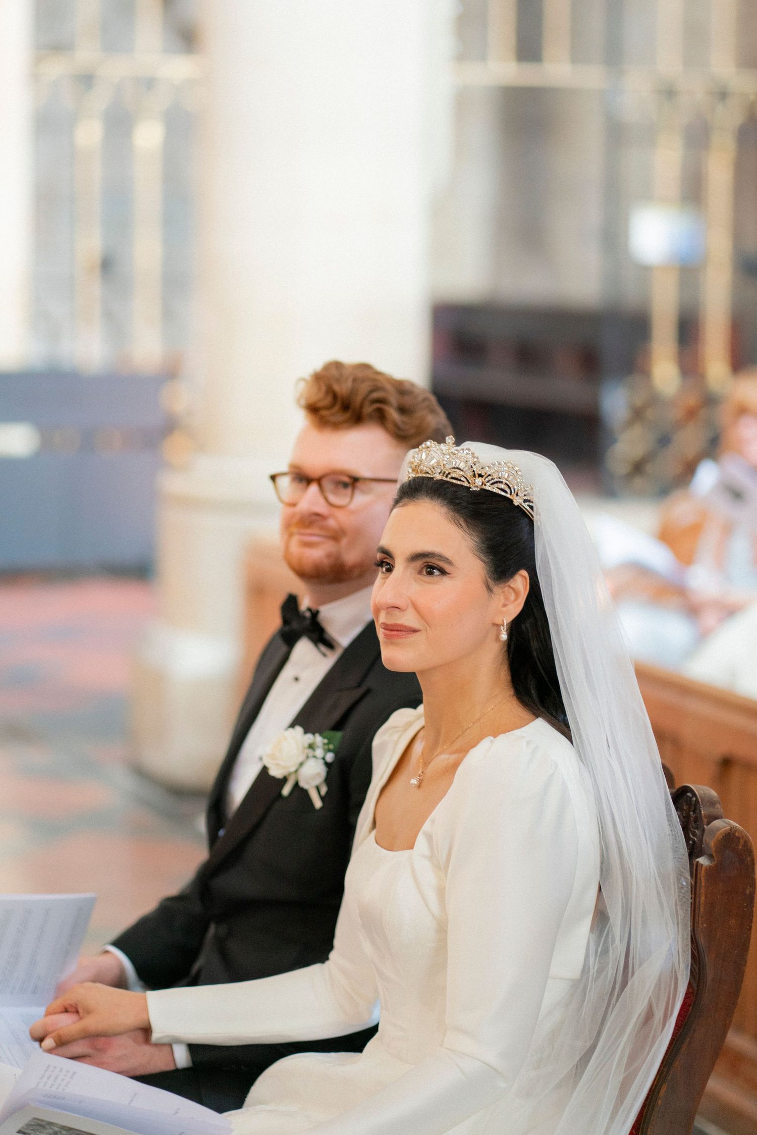 Bride in tiara and veil sits with groom in tuxedo during wedding ceremony in elegant church.