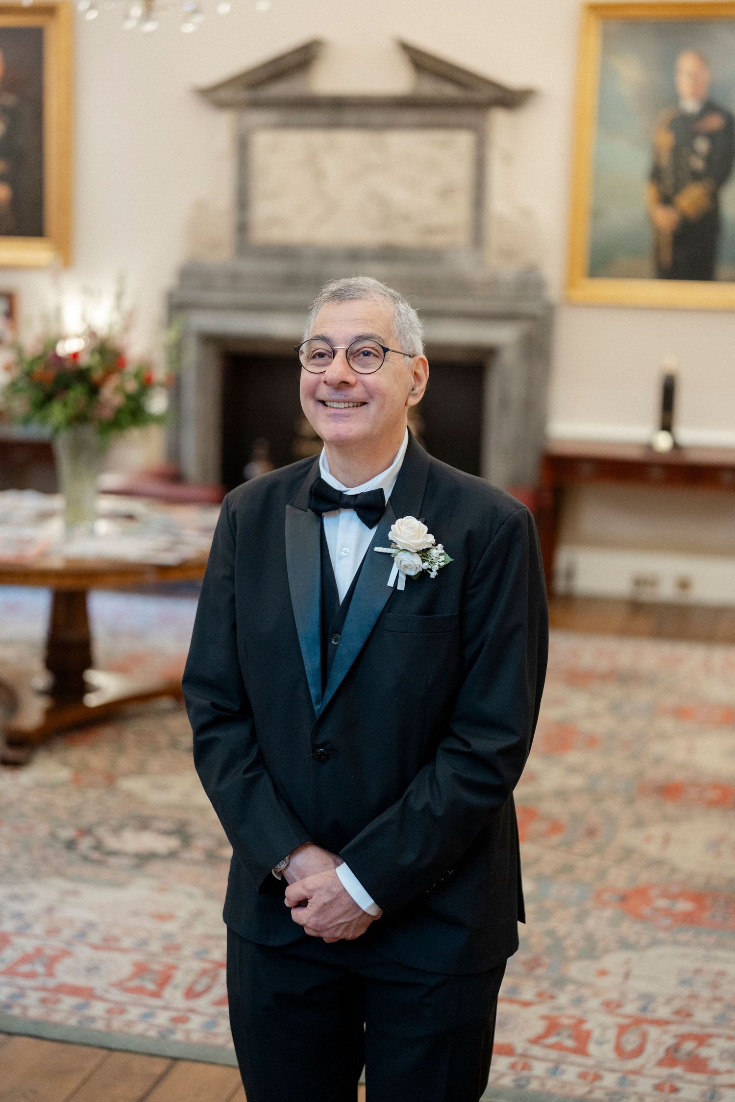 Smiling groom in black tuxedo with white boutonniere stands in elegant room with portraits and fireplace.