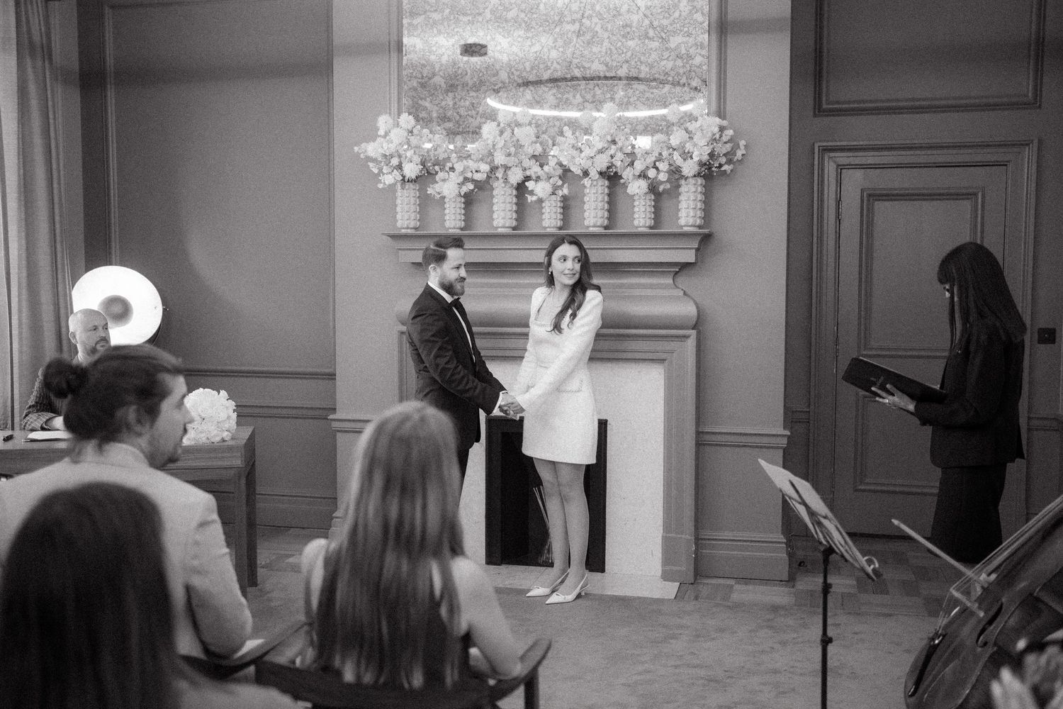 Black and white photo of a couple exchanging vows at an intimate indoor wedding ceremony before a fireplace.