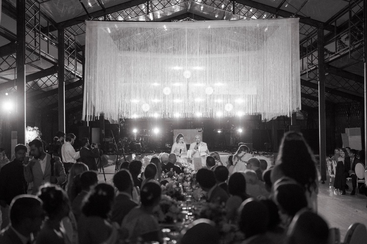 Black and white wide shot of elegant wedding reception in industrial venue with draped backdrop and chandelier.