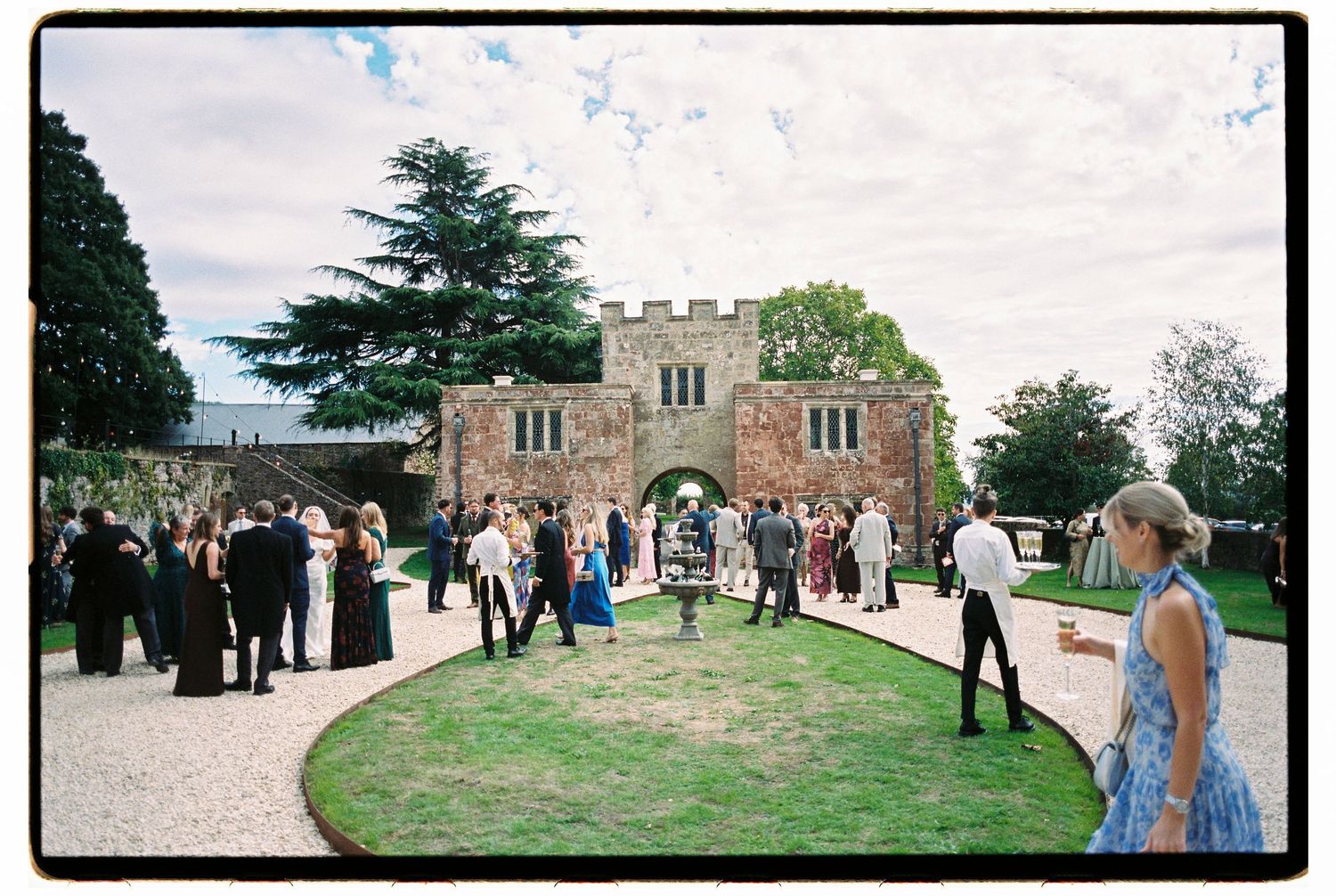 Elegant outdoor wedding reception at a historic stone gatehouse venue with guests mingling on manicured grounds.