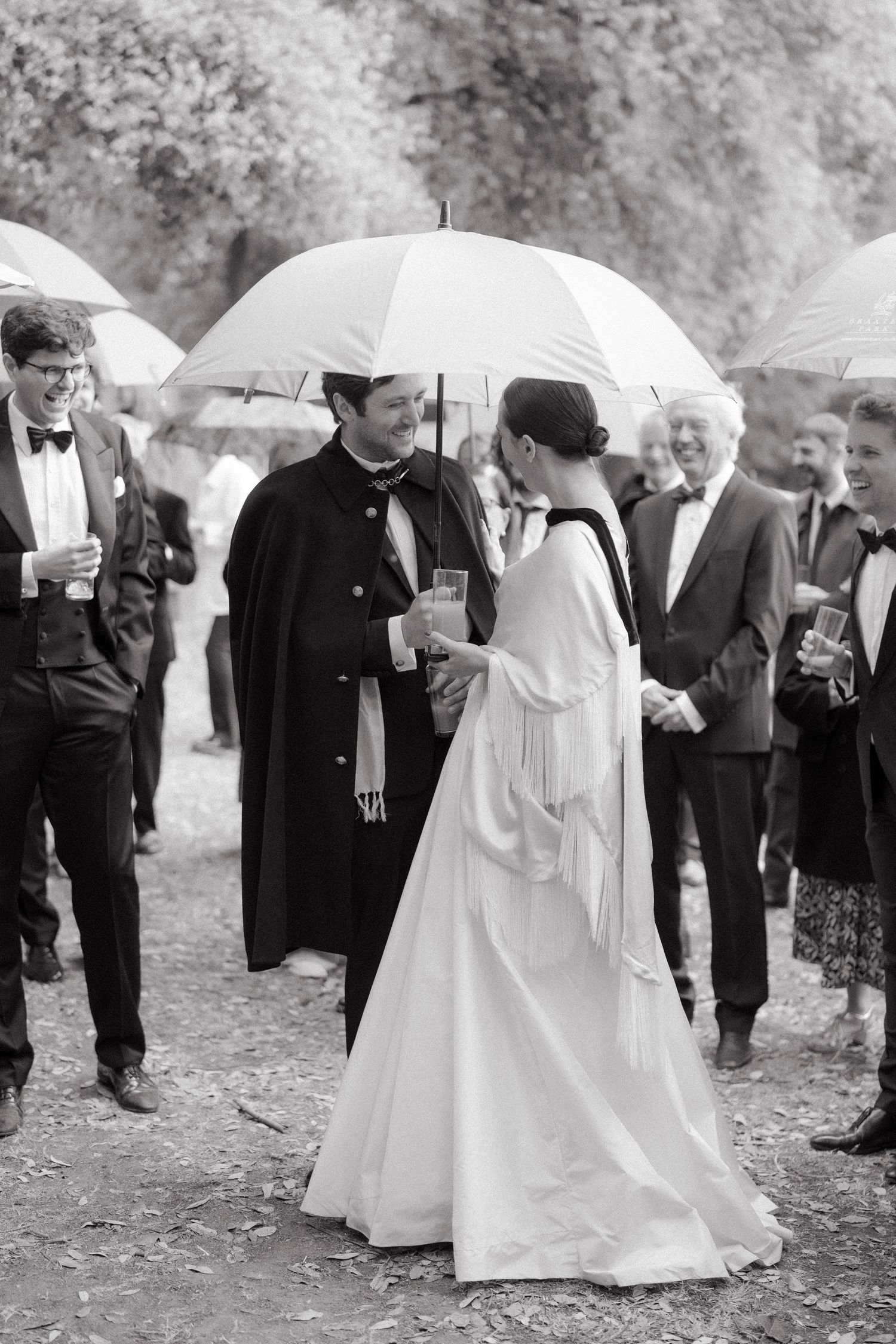 Black and white photo of a bride in a flowing gown and a groom sharing a white umbrella as formally dressed wedding guests chat outdoors.