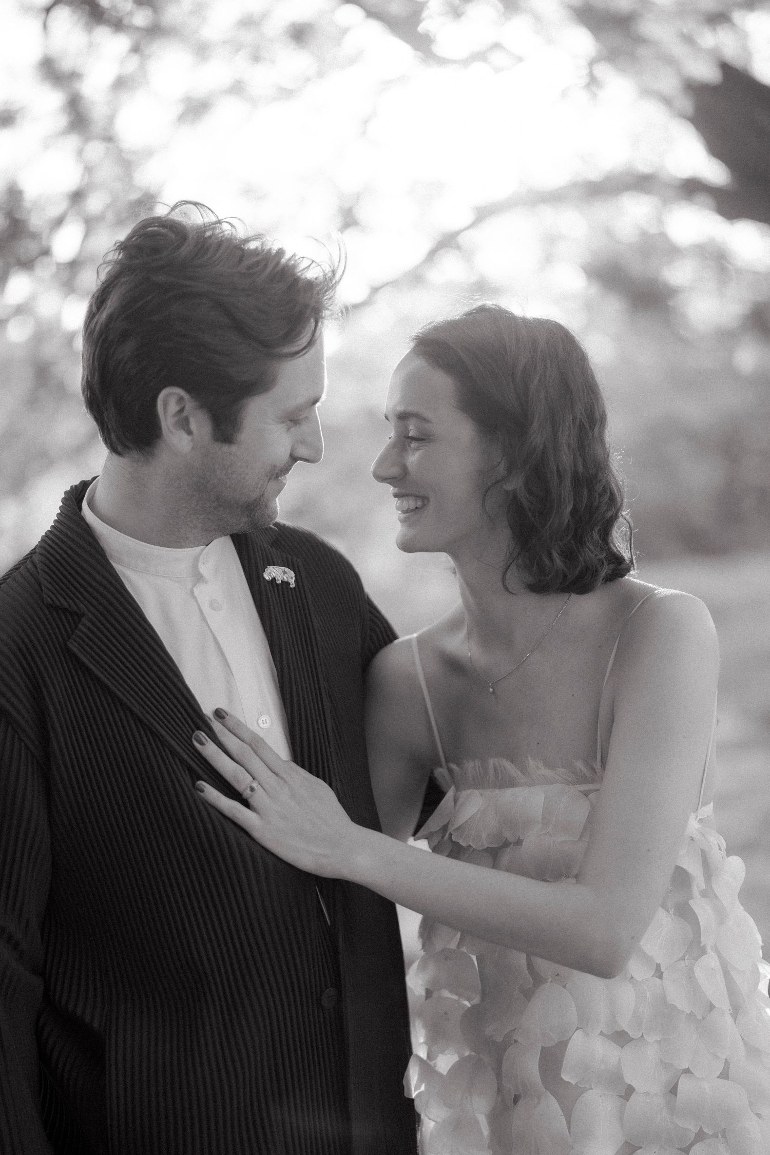 A smiling couple shares a tender moment outdoors in a black and white portrait, her hand resting on his jacket lapel.