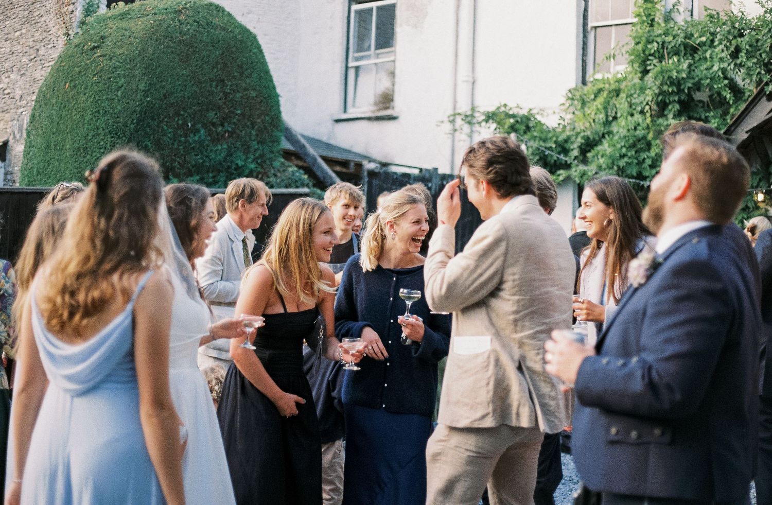A group of elegantly dressed guests socializing at an outdoor garden party with drinks in hand.