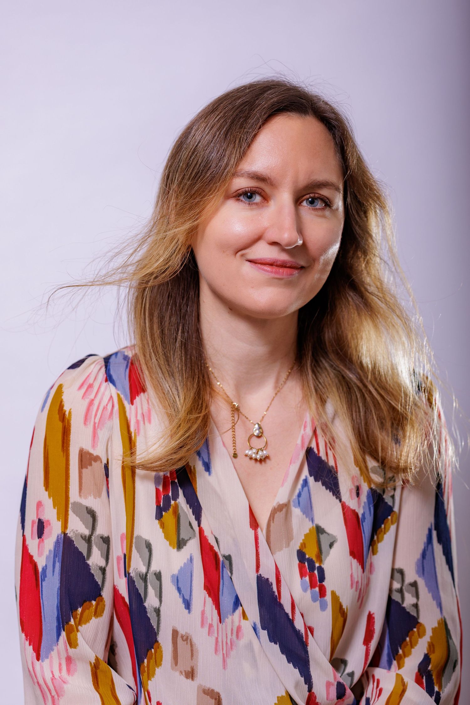Smiling woman with long blonde hair wearing a colorful patterned blouse against a grey background.