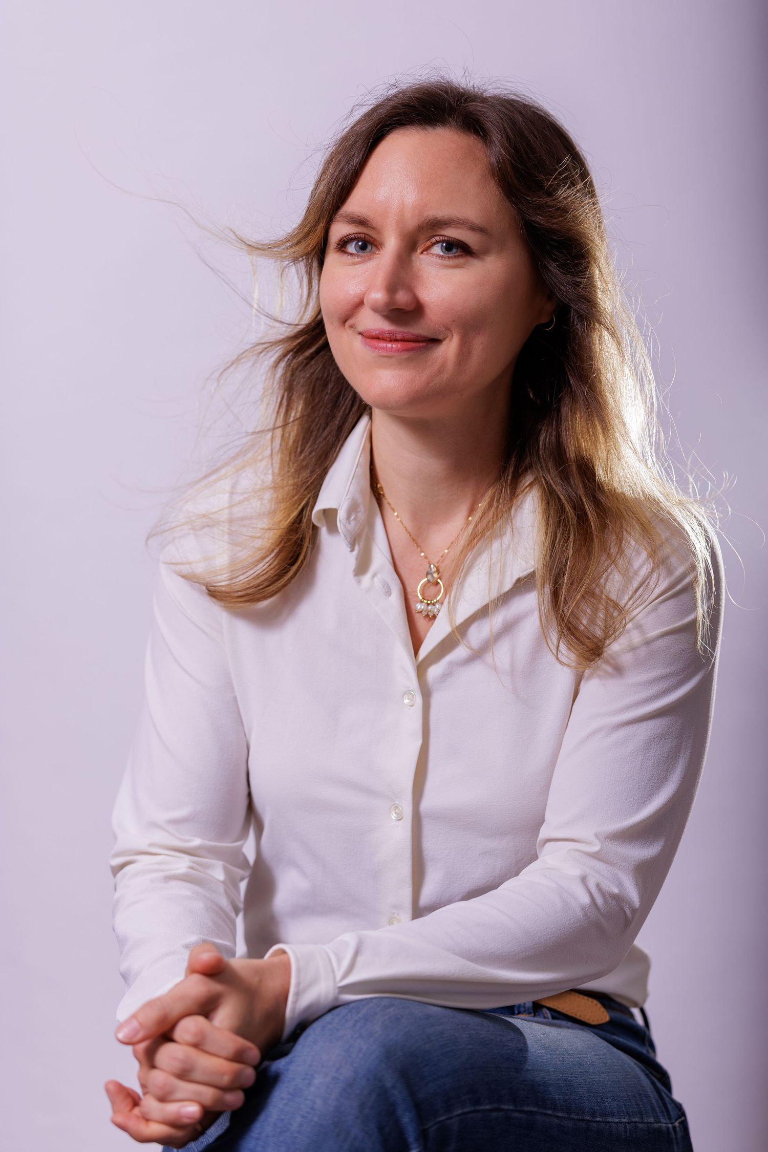 Smiling woman with long blonde hair in a white shirt and jeans seated against a light grey background.