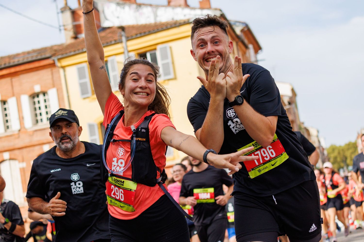 Two marathon runners celebrate joyfully at a race finish line wearing numbered bibs in a sunny outdoor setting.