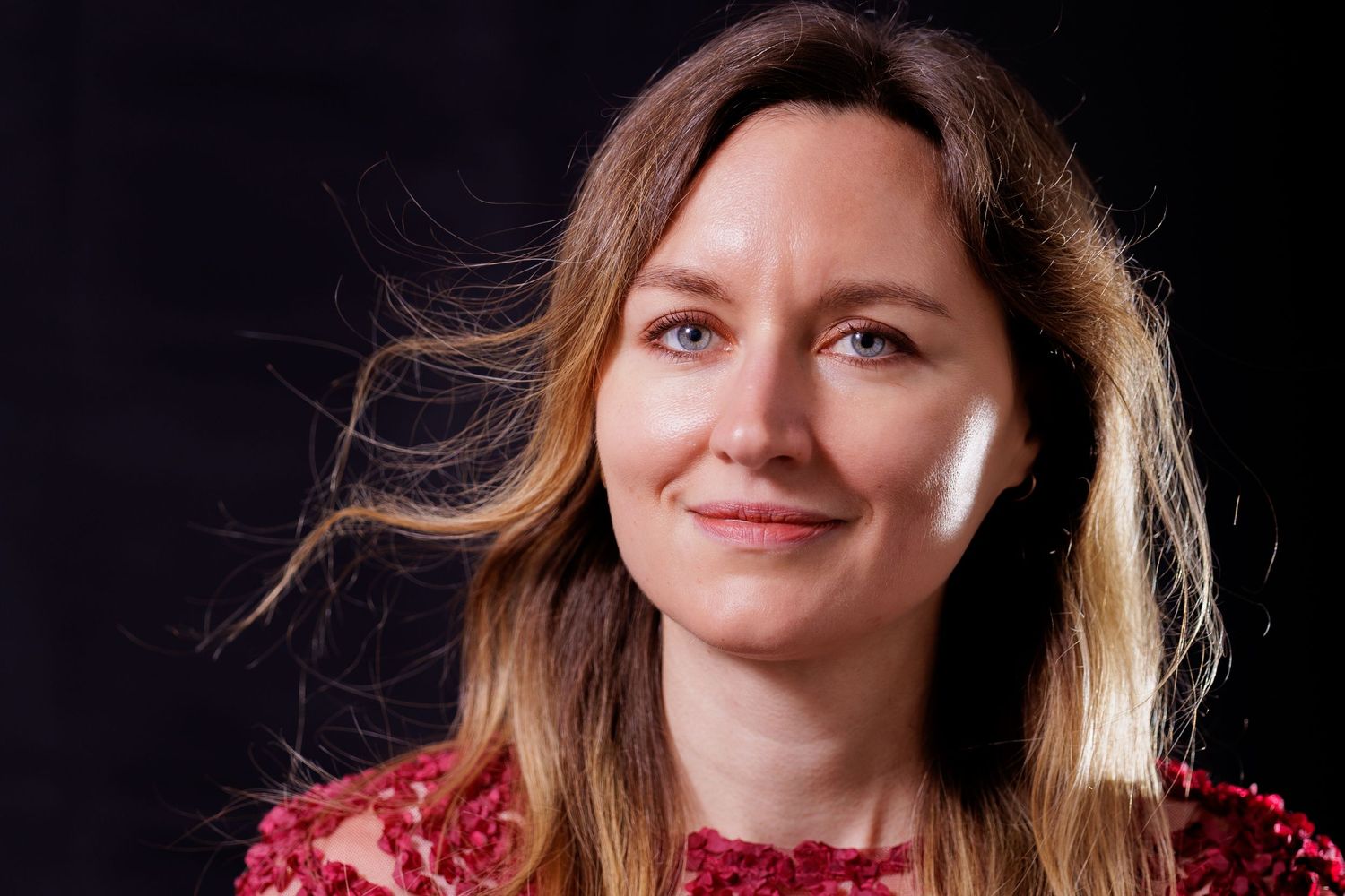 Smiling woman with long brown hair wearing a red floral top against a dark background.