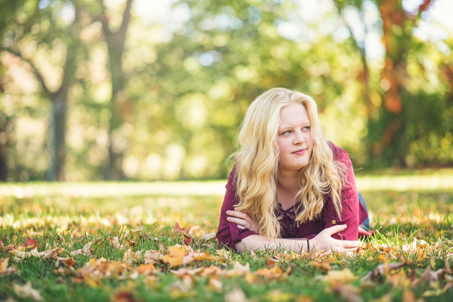 Person in burgundy sweater lying on colorful autumn leaves during outdoor portrait session.