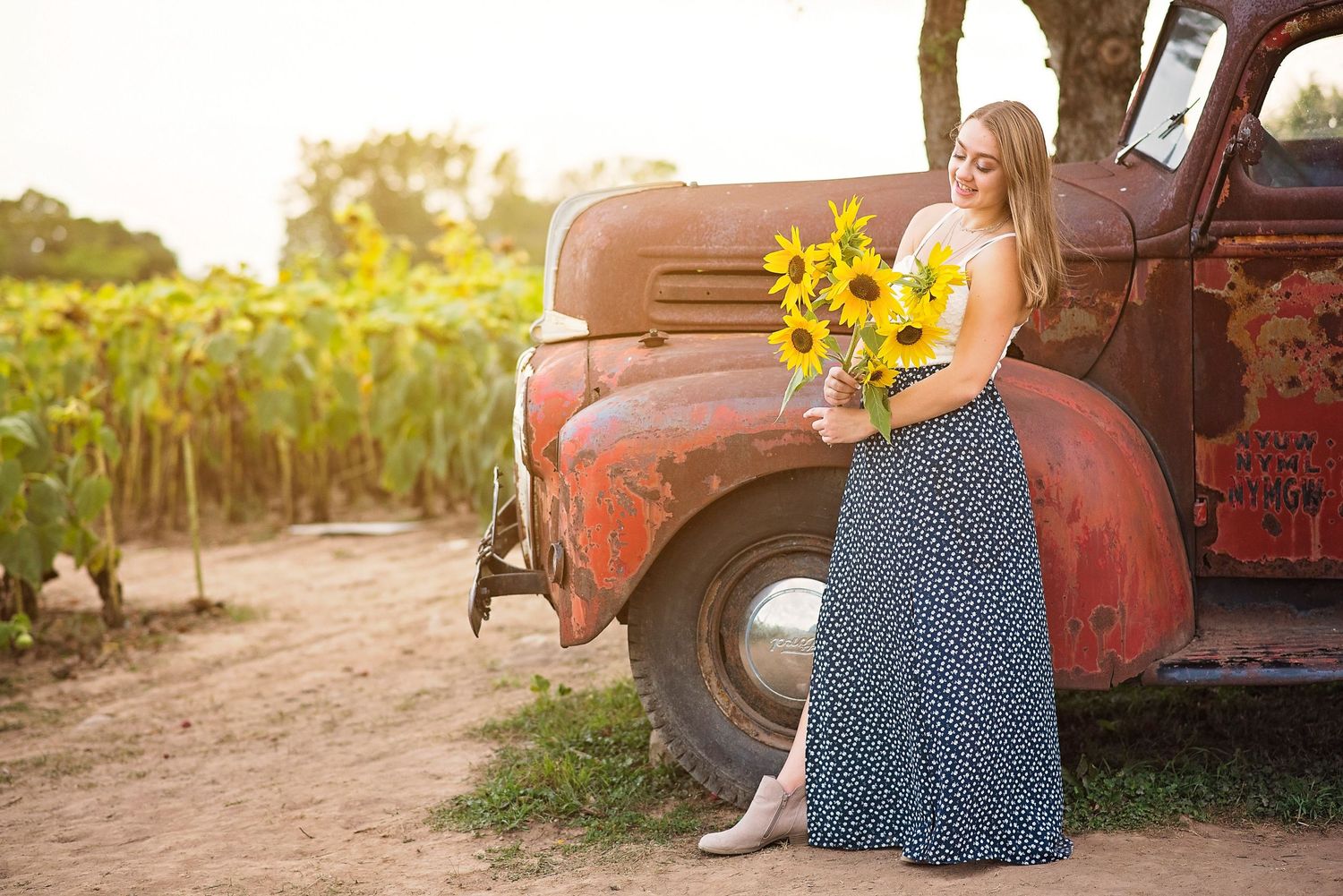 A person in a flowing skirt poses with sunflowers next to a vintage red truck at sunset.