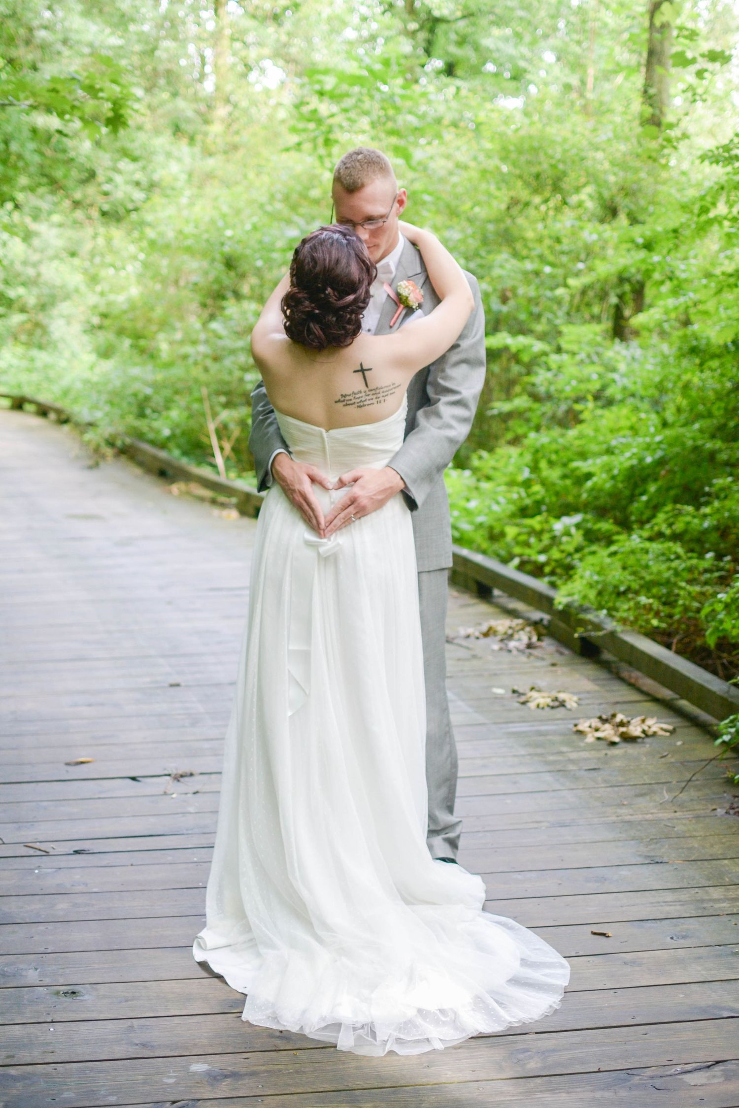Newlyweds embrace on wooden walkway surrounded by green forest.
