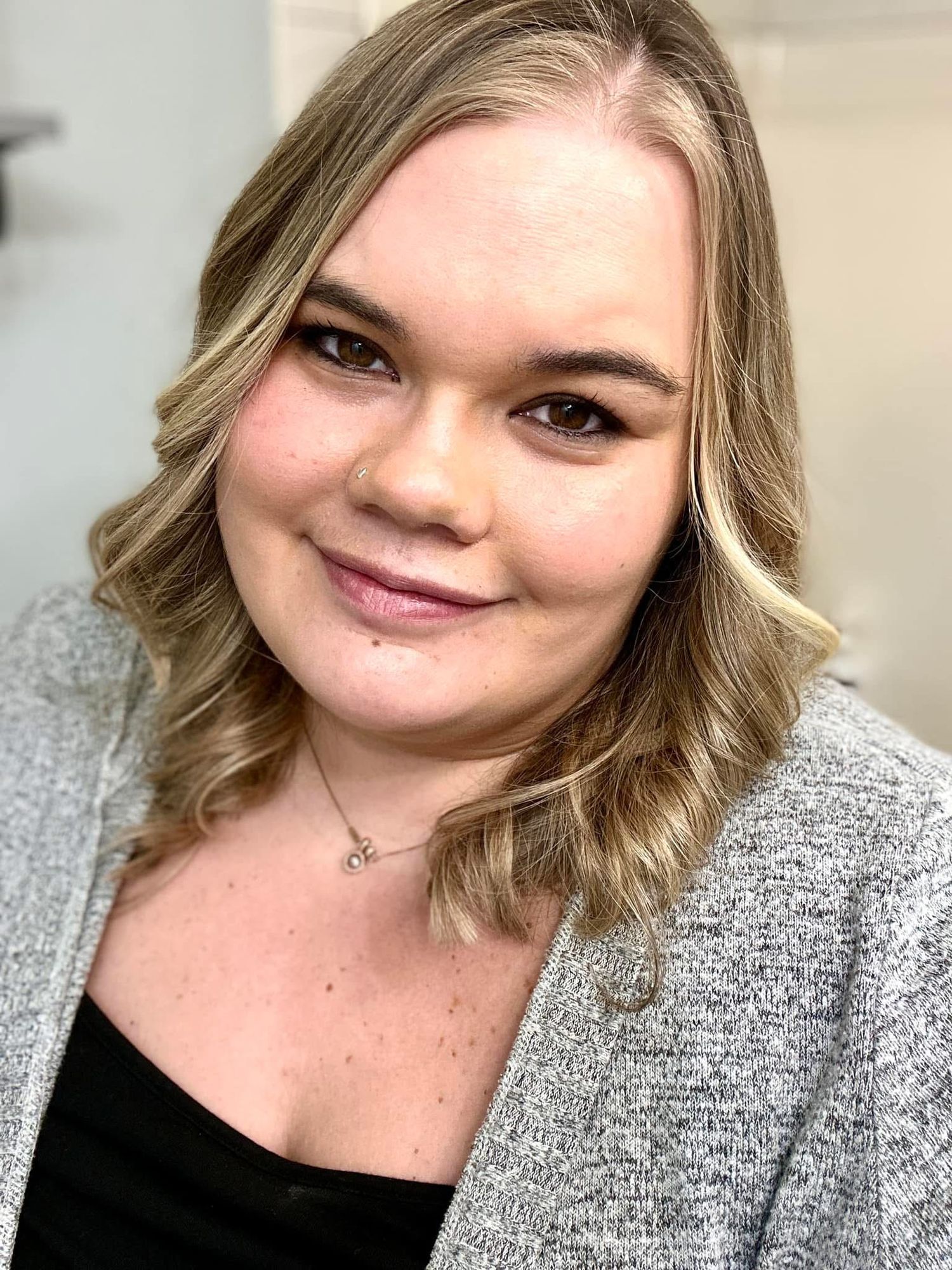Professional headshot of a person wearing a gray cardigan and black top with styled blonde waves.