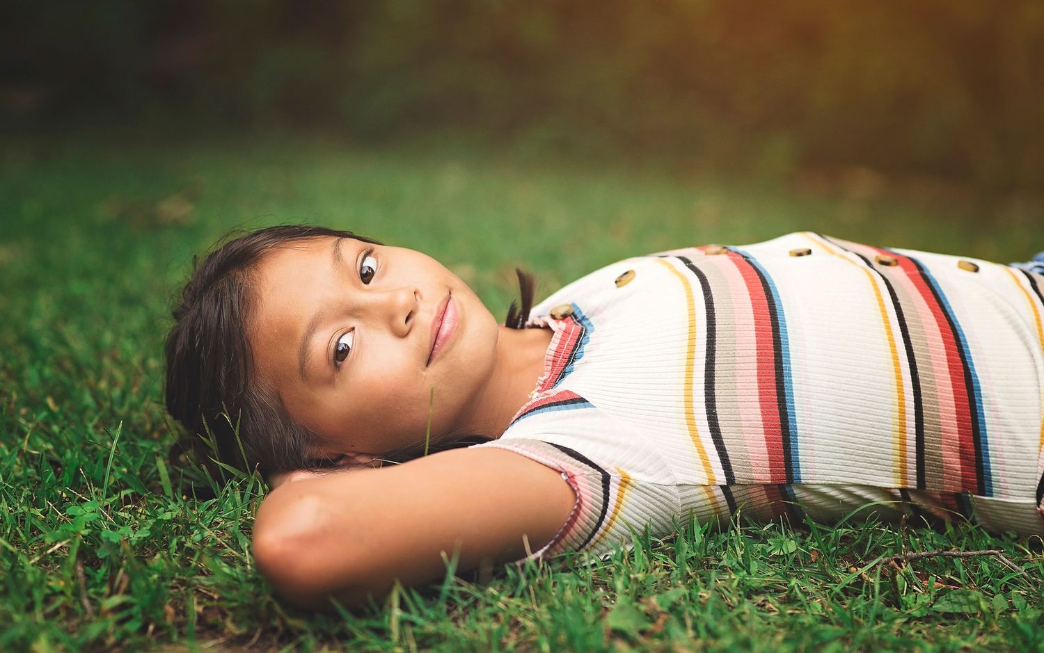 Young person lies peacefully on grass wearing striped shirt on summer day.