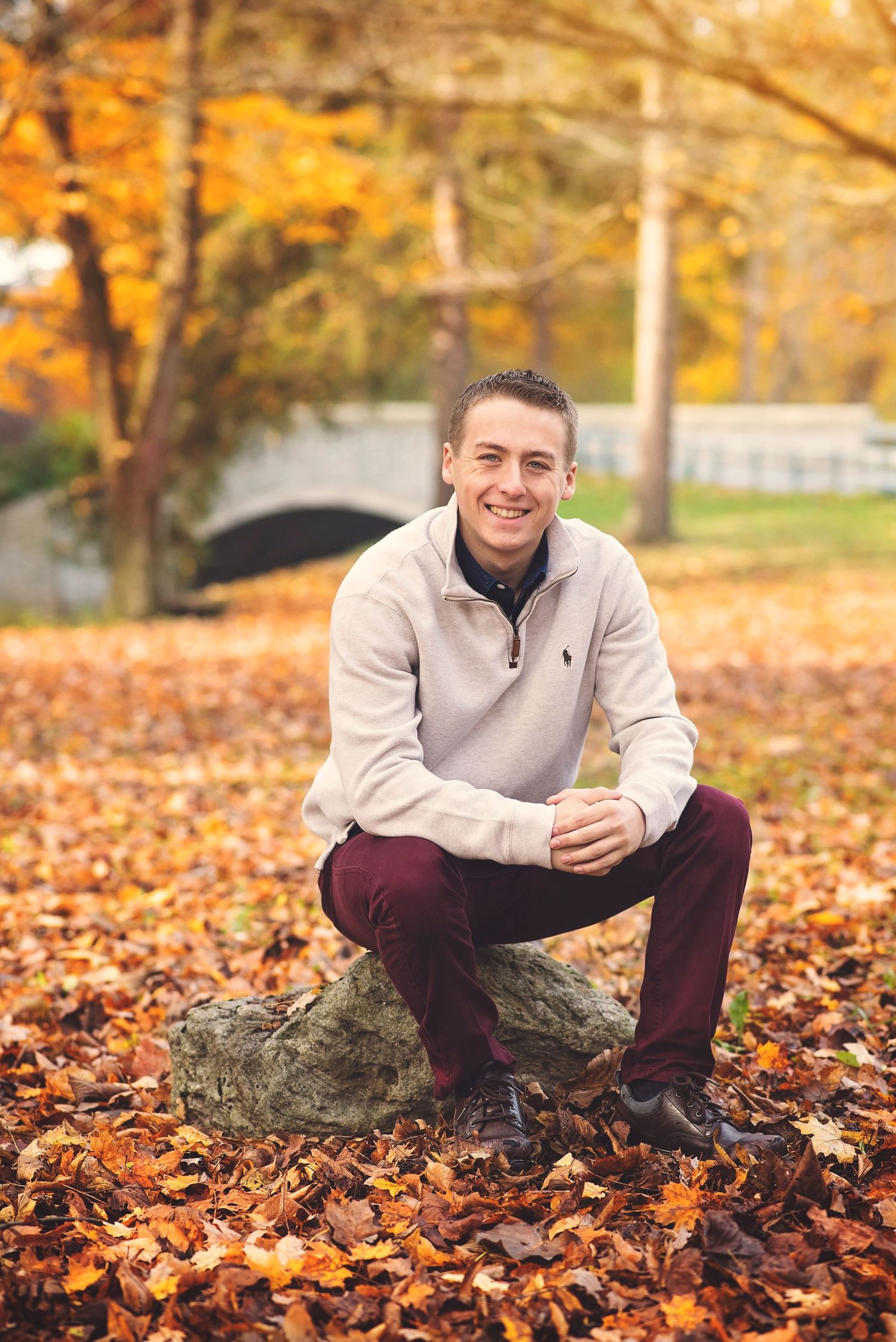 Person in beige quarter-zip sweater sitting on rock surrounded by autumn leaves.