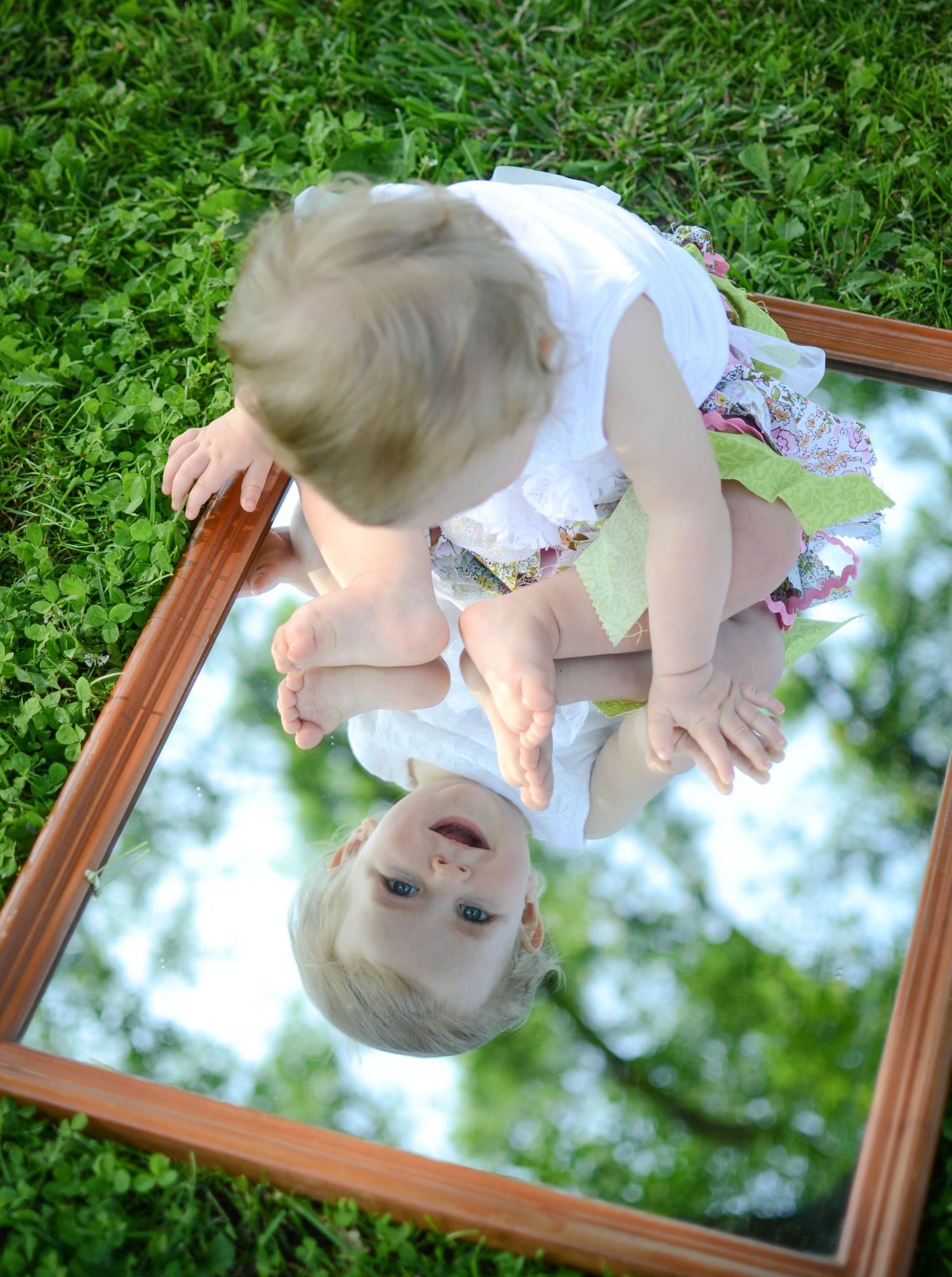 Creative overhead shot of baby in white dress reflected in mirror on grass.