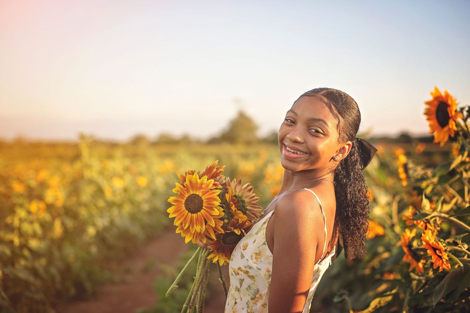Person holding sunflowers smiles while posing in a white dress during sunset.