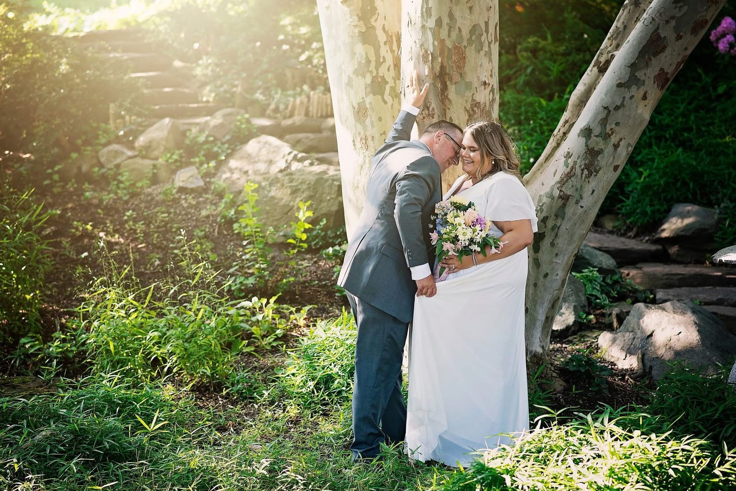Wedding photograph of a couple embracing under a tree with sunlight filtering through the branches.