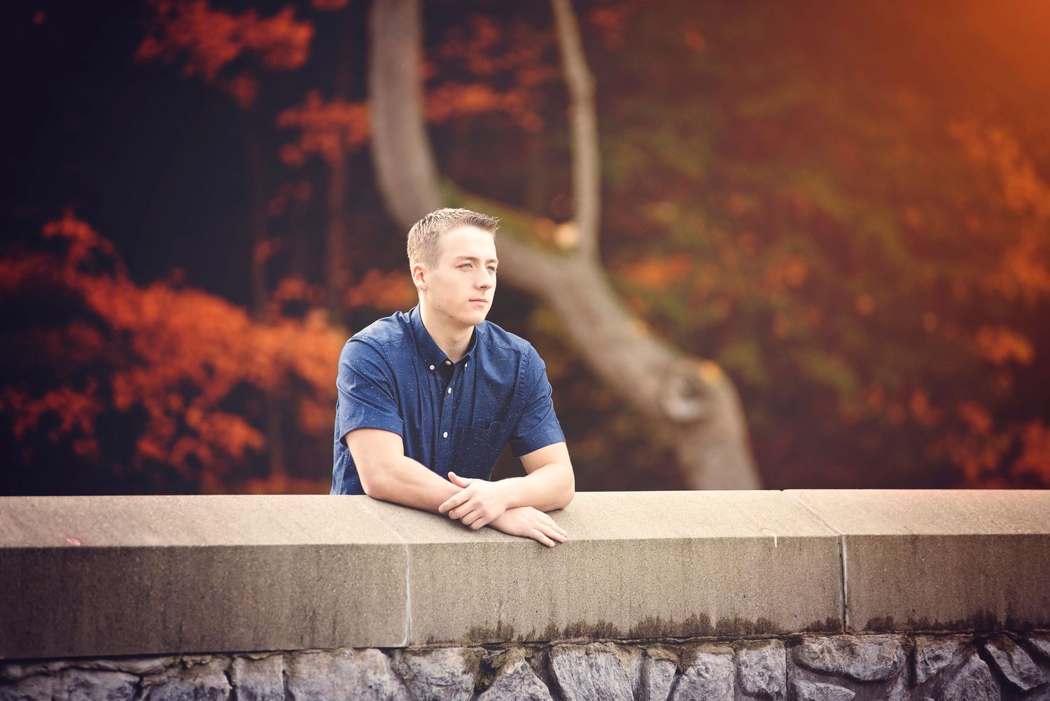 Person in blue shirt leaning on stone wall with autumn foliage in background.