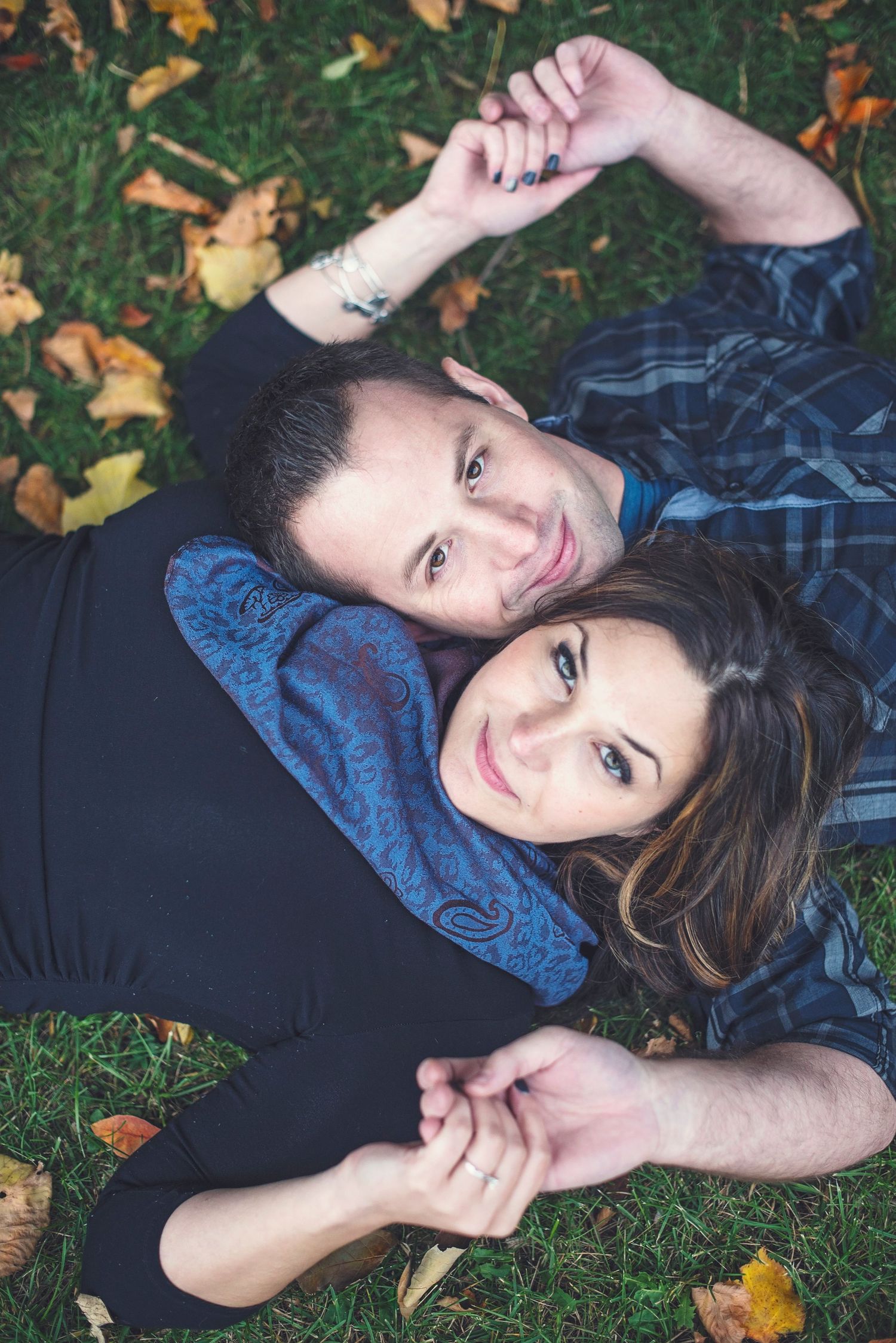 A couple lying on autumn leaves in casual attire smiling at the camera while holding hands.