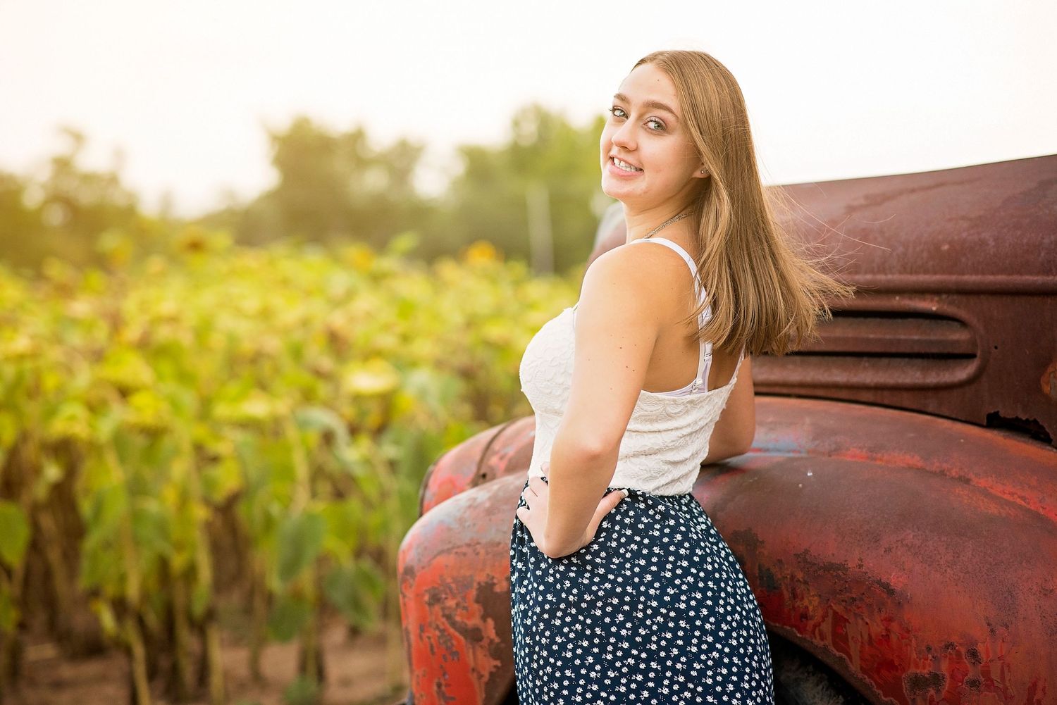 A person in a patterned skirt leans against an old red truck in a sunflower field.