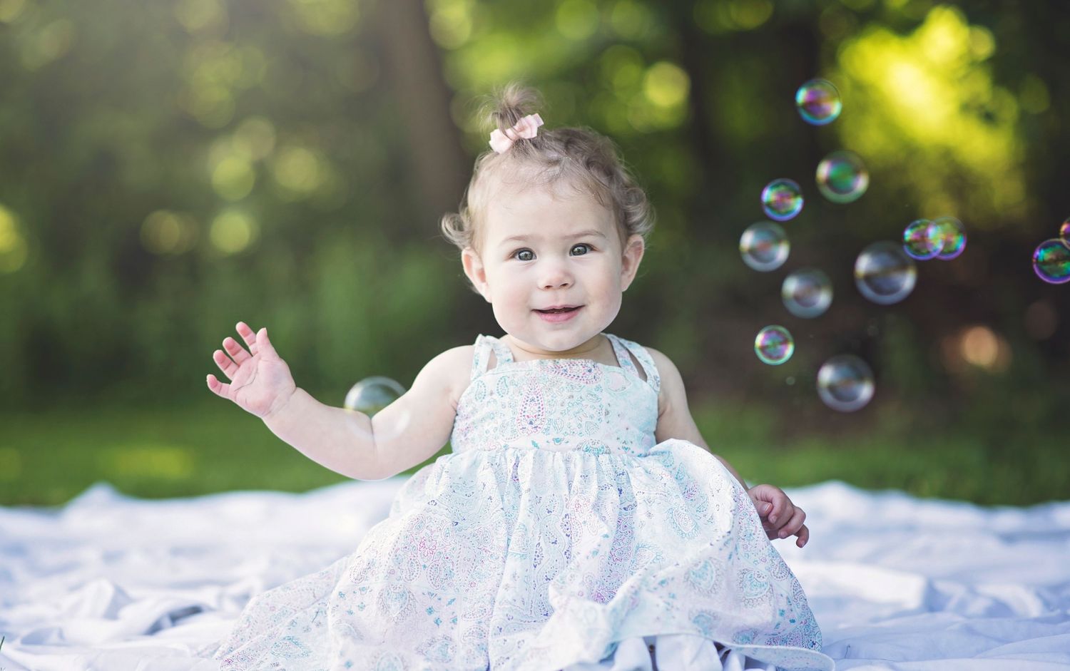 Baby in sparkly dress playing with bubbles on white blanket outdoors.