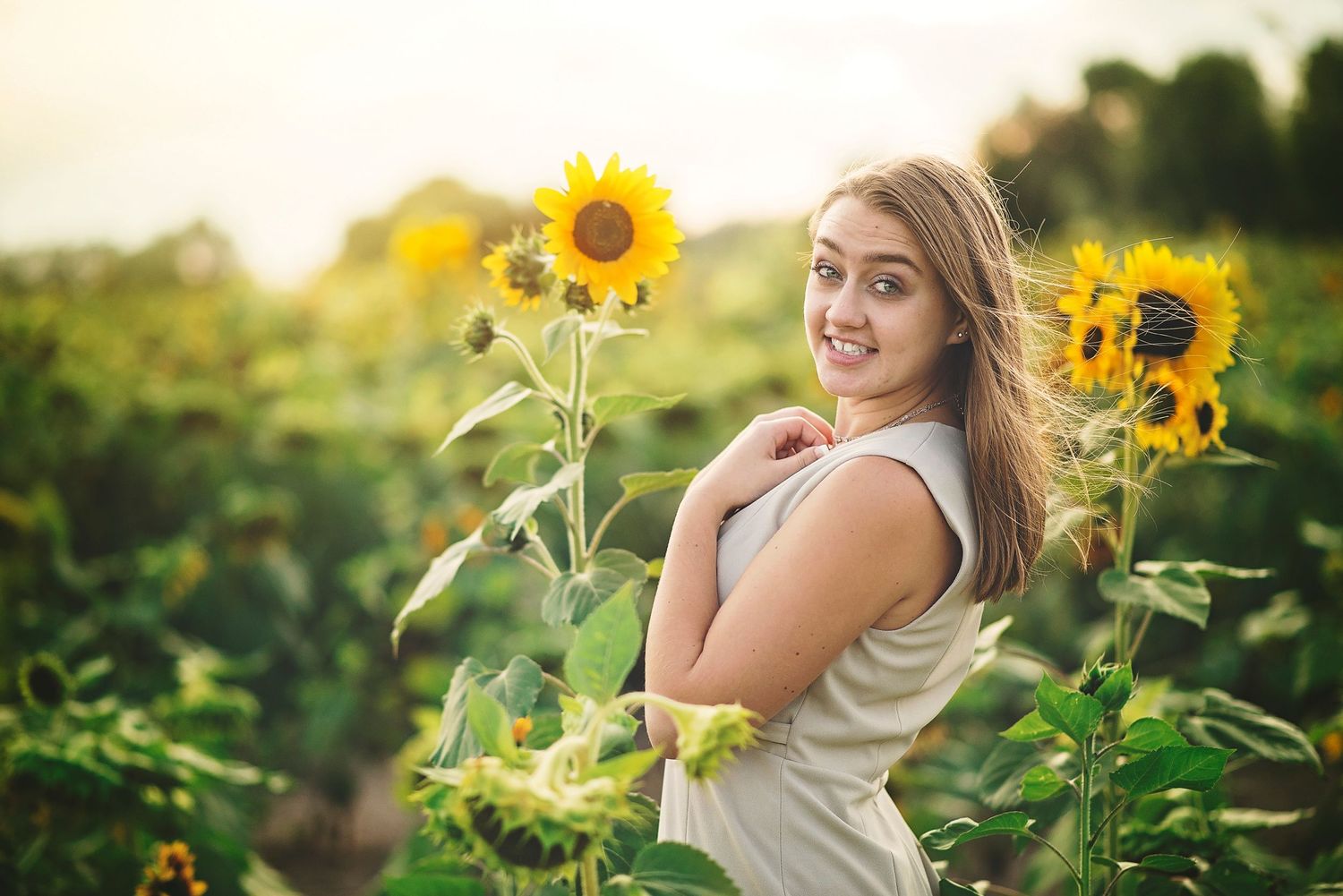A person in a white top poses playfully among tall sunflowers in a garden setting.