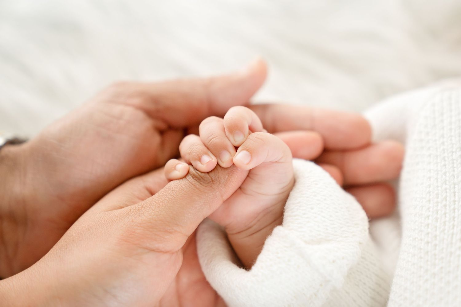 A tiny newborn baby hand grasps a parent's finger while wrapped in soft white blanket.