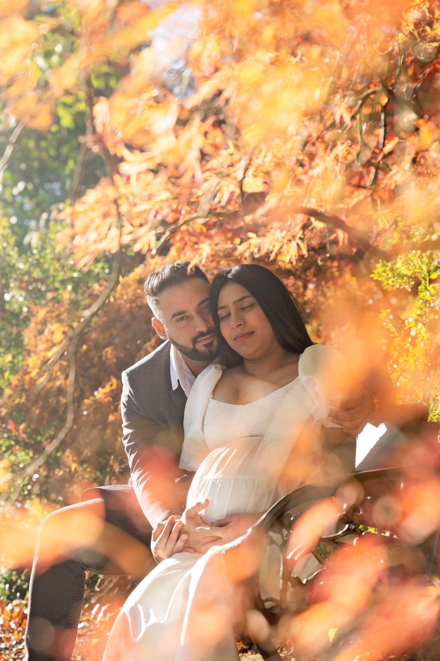 A romantic couple embraces beneath vibrant orange autumn foliage in a glowing sunlit garden setting.
