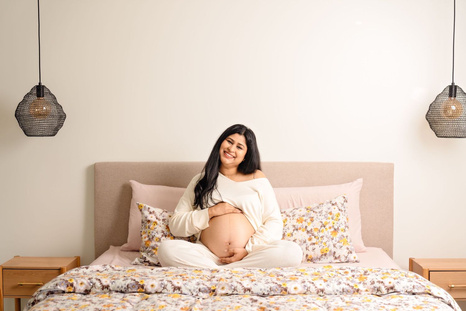 Expectant woman in white sweater relaxes on bed with floral bedding and geometric pendant lights overhead.