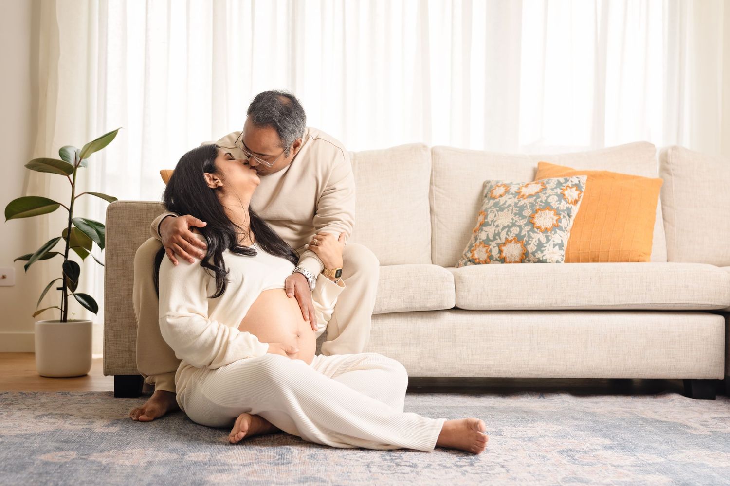 A couple in cozy neutral outfits relaxes together on a plush white sofa in a bright, modern living space.
