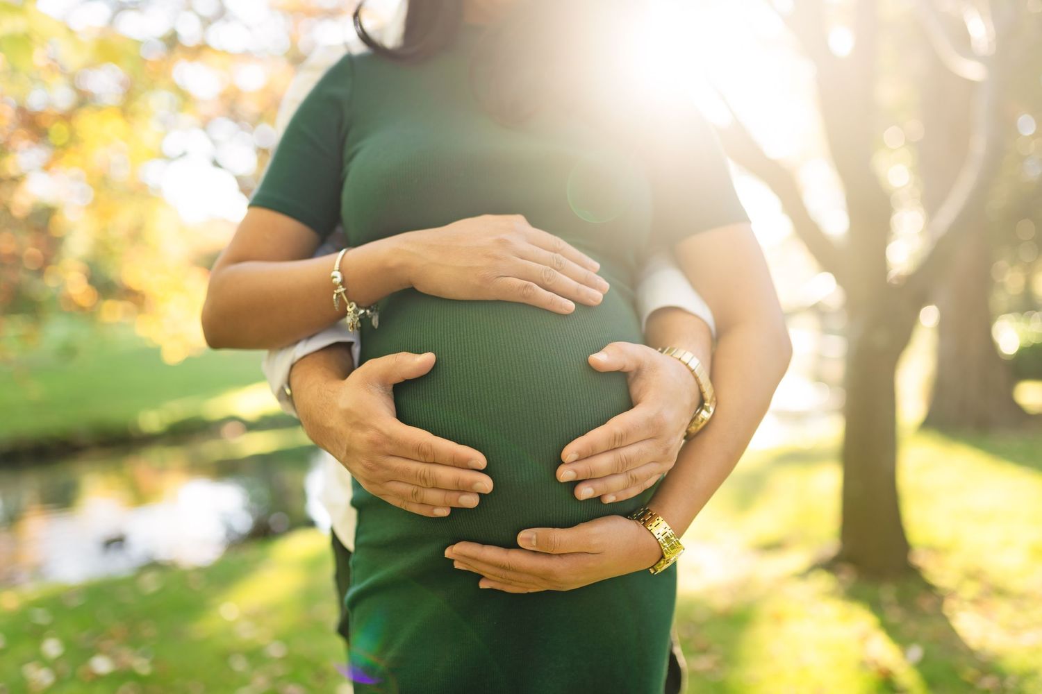 A pregnant woman in a green dress cradles her belly while standing in a sunlit park during golden hour.