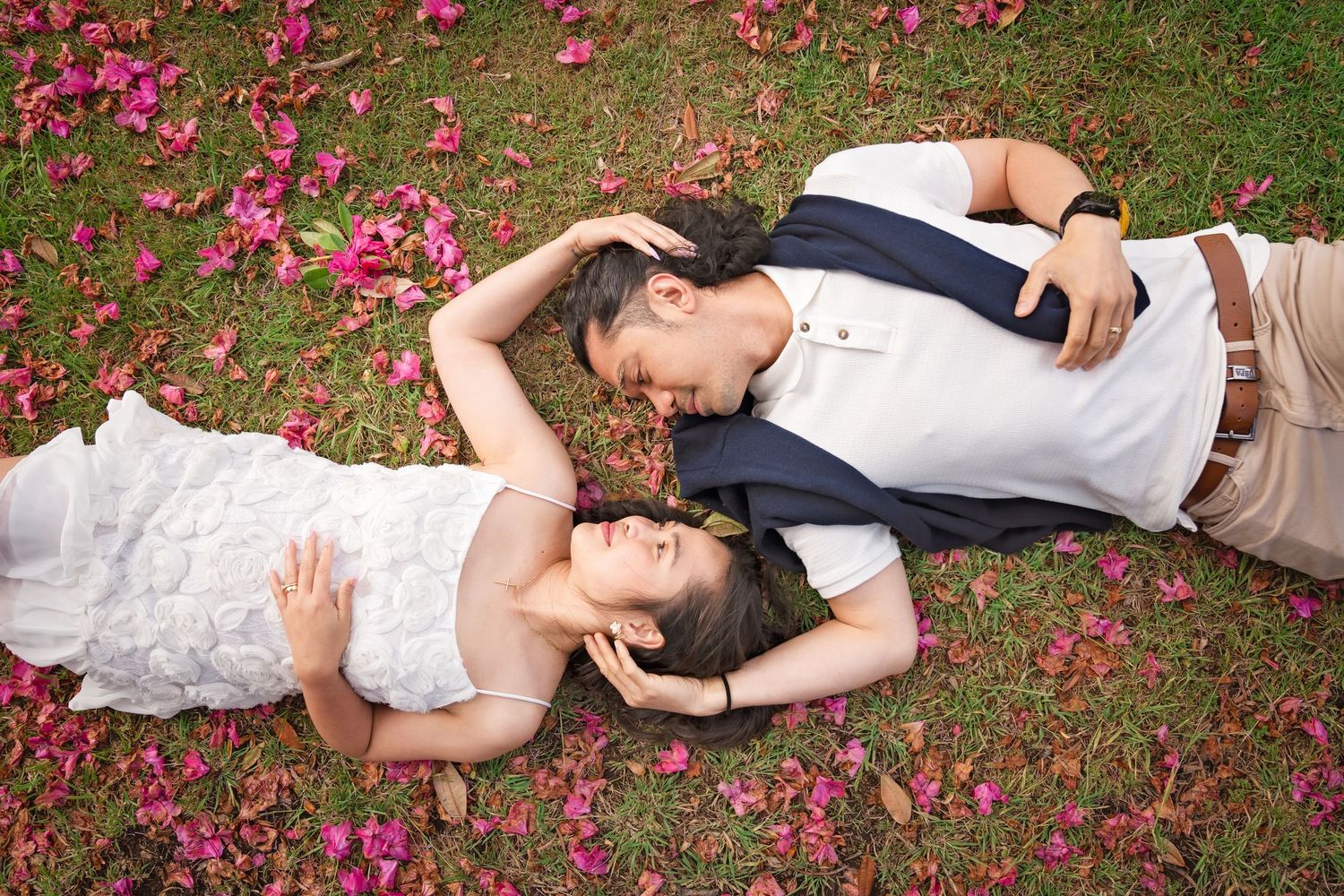 Romantic couple lying on grass surrounded by scattered pink flower petals in a garden.
