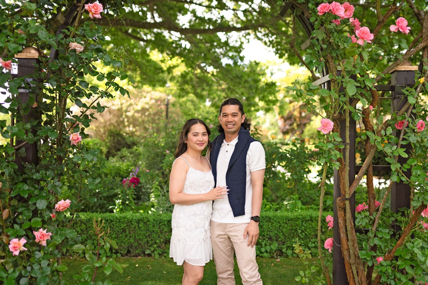 Couple smiling under floral arch in lush garden with pink roses.